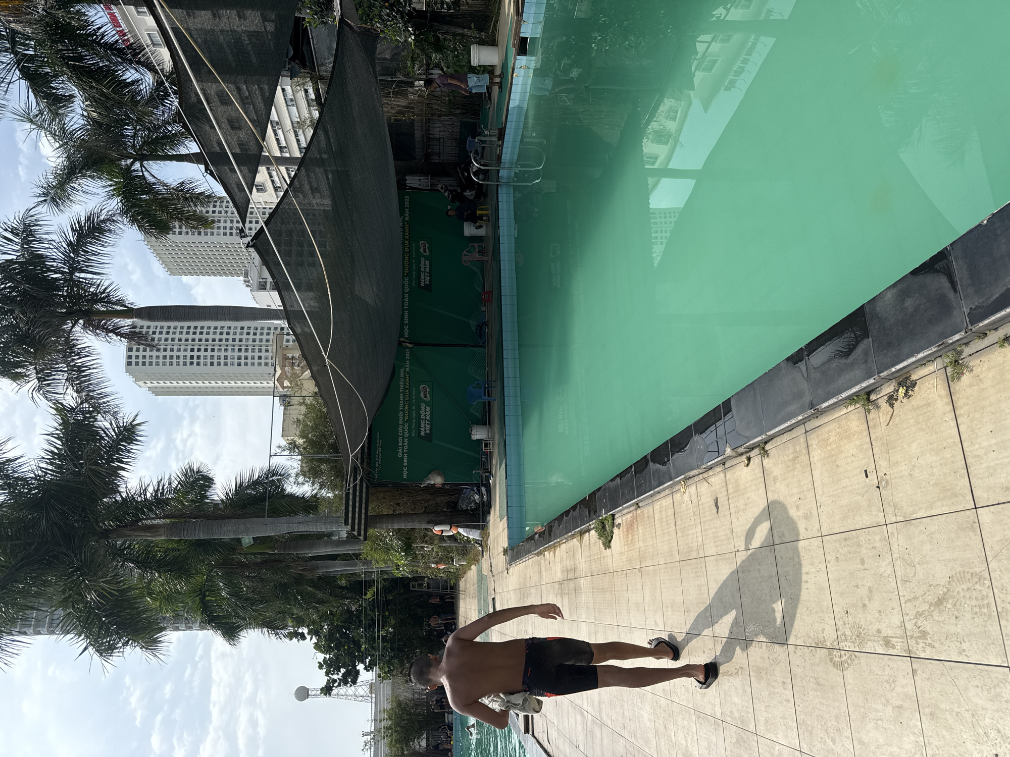 The image depicts an outdoor swimming pool scene in Nha Trang, Vietnam. A young male, partially undressed and carrying a towel, walks away from the camera on a tiled path alongside the bright turquoise water of the pool. The pool area is shaded by a dark mesh canopy, and tall palm trees are visible, along with the white facades of high-rise buildings in the background, suggesting an urban setting. A green banner with Vietnamese text and logos, including "Milo," is stretched across a fence behind the pool. Several other individuals are present in the background, some near the pool and others walking around. The overall environment suggests a public or shared swimming facility.
