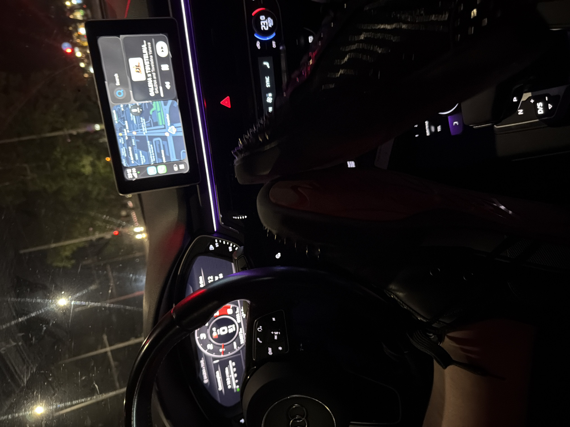 The image presents the interior of a dark Audi vehicle at night, observed from a low angle. The vehicle appears stationary or moving slowly, indicated by a 0 km/h reading on the digital instrument cluster, which also displays an odometer reading of 109,727 km.
On the left, a black multi-function steering wheel with the Audi emblem is prominent. A leg with a black laced shoe is partially visible in the lower left, positioned near the implied pedal area. The dashboard features a purple ambient light strip.
A central infotainment screen is active, displaying Apple CarPlay. The left portion shows a navigation map, while the right displays a media player. The media player shows the song "GALENA X TSVETELINA..." by "GALENA and Tsvetelina Yaneva," suggesting a Bulgarian origin for the music. A faint "Ruse" suggestion is discernible in the search bar area of the screen, indicating the vehicle's location is in the city of Ruse, Bulgaria.
To the right, a pair of legs is elevated, with the right foot resting on the dashboard above the climate control panel, which is set to "23.0 AUTO." This foot wears a black, high-heeled shoe adorned with studs or spikes and a distinct red sole, characteristic of Christian Louboutin footwear. The hazard lights (red triangle) are illuminated on the center console.
Beyond the windshield, blurred streetlights, a red traffic light, and a dark urban environment are visible, along with streaks on the glass.