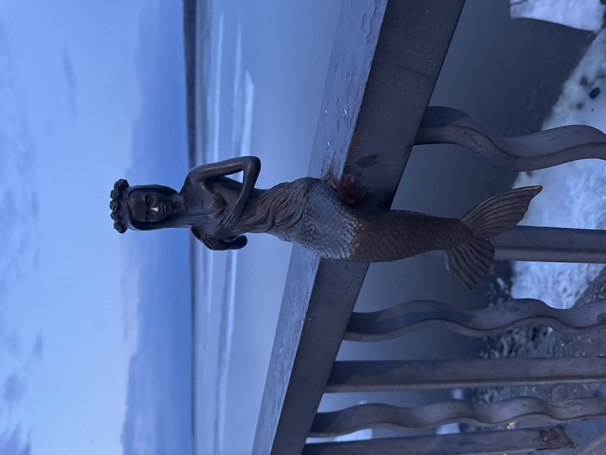 The image displays a dark-colored statue of a mermaid integrated into a metallic railing, set against a backdrop of water and sky.
The central subject is a mermaid statue, likely cast in bronze or a dark metal, positioned upright. She wears an ornate crown or elaborate hairpiece and has a human upper body with arms bent, hands held near her torso. Her lower half transitions into a textured fish tail, which forms part of the decorative railing structure.
The railing itself is dark and robust, featuring a thick, flat top bar. Beneath this, several wavy horizontal bars create an artistic pattern, evocative of ocean waves or currents. The railing exhibits signs of weathering, with some visible rust or discoloration. To the right, at the base of the railing, there are hints of snow or ice on the ground, suggesting a cold environment.
In the background, a wide expanse of calm, blue-grey water stretches to the horizon, indicative of a large lake or river. Above, the sky is a soft, muted blue, largely covered by clouds, which contributes to the dim, diffused lighting. The overall scene suggests either twilight (dusk or dawn) or a heavily overcast day.
The image captures a static scene with no discernible activity or events occurring. There is no visible text within the frame. The context provided indicates the location is Chaikovsky, Russia.