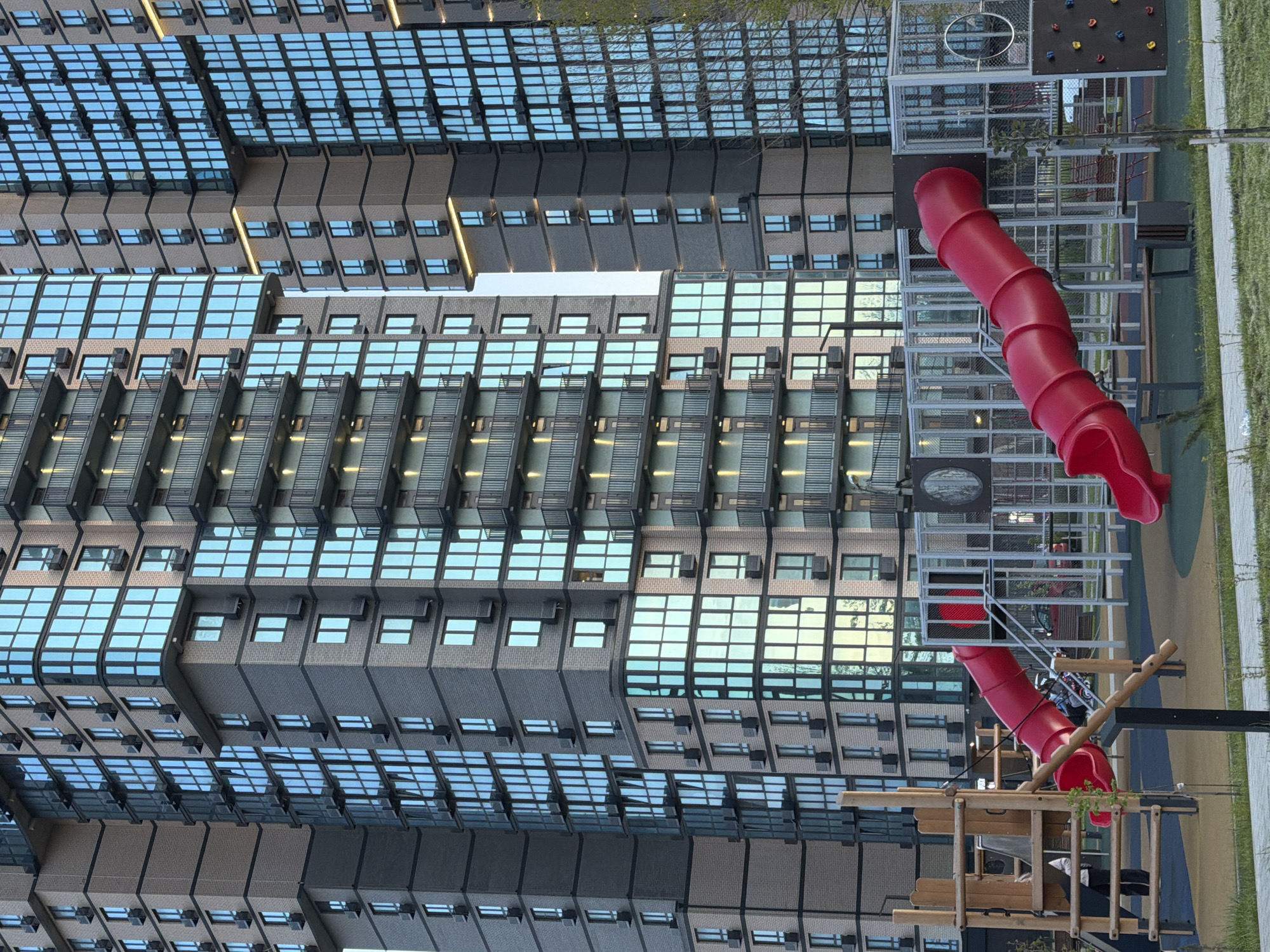 The image captures a modern urban residential landscape featuring several tall, contemporary apartment buildings and a children's playground. The buildings, which dominate the left and central parts of the frame, display a distinctive architectural design with repetitive patterns. Their facades are composed of light brown, brick-like segments interspersed with large, rectangular glass windows, many of which reflect the clear sky and surrounding environment. Some sections feature balcony-like structures or recessed areas with horizontal slatted elements.
In the foreground and to the right, a colorful children's playground provides a recreational space. It includes two bright red tubular slides: a prominent, winding one supported by a multi-tiered metal framework with stairs and platforms, and a smaller one. Additional playground features visible are a small rock climbing wall with various colored holds, a circular metal ring, and a wooden play structure resembling a fort or bridge. The ground in the playground area is covered with green artificial turf or grass. Sparse greenery, possibly small trees or bushes, can be seen integrated within the scene.
The photograph appears to be taken during the daytime, under clear weather conditions, with the lighting suggesting either morning or late afternoon. A single person is partially visible near the wooden playground structure, appearing to be sitting or resting. The scene conveys the orderly environment of a modern residential development. There is no visible text in the image.