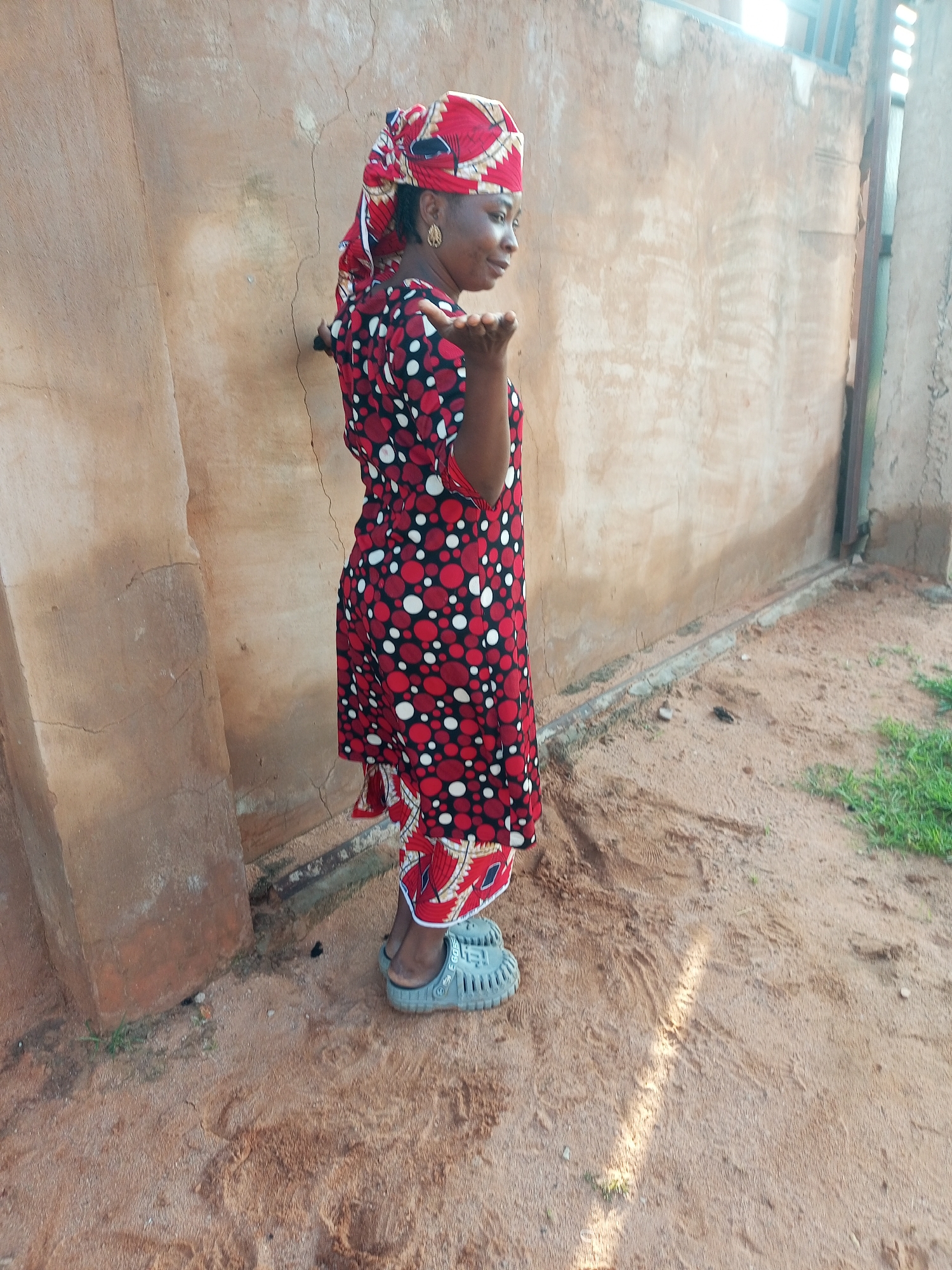 A woman stands outdoors, facing right, in the city of Tempure, Nigeria. She wears a red headscarf with a white and black circular pattern, a red dress with a similar pattern, and light grey clogs. Her right hand is raised, palm facing outward, as if blowing a kiss. The background consists of a rough, tan-colored wall and sandy ground with sparse patches of green grass. The lighting suggests it is daytime, with sunlight casting shadows and highlights.
