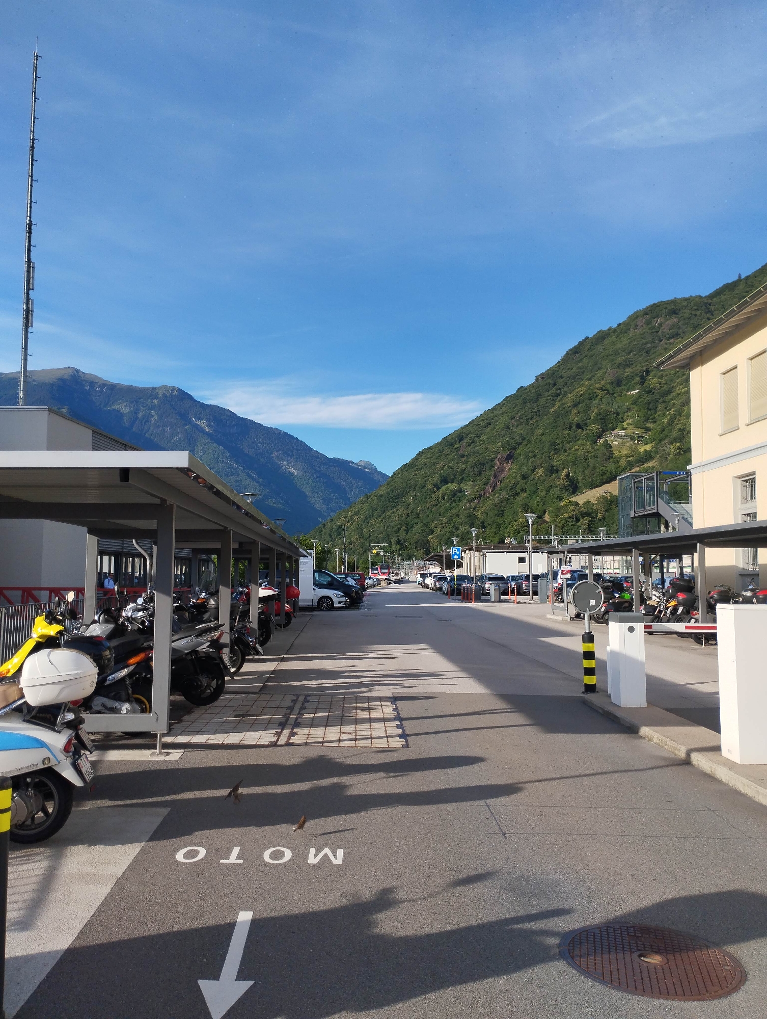 The image depicts an outdoor scene dominated by mountainous terrain under a clear blue sky. In the foreground, an asphalt road features painted markings: a large white arrow pointing downwards and the word "MOTO" (appearing upside down from the viewer's perspective). Several small, dark birds are perched on the asphalt near the painted word. To the left, a long, covered parking structure shelters numerous motorcycles and scooters, including a prominent yellow one on the far left. A tall, slender telecommunications tower stands against the sky in the left background.
On the right side of the road, another similar covered parking area is visible, with more motorcycles parked beneath it. Beyond this, a multi-story, light-colored building with rectangular windows stands next to a street lined with parked cars, extending into the distance. The background is dominated by imposing, steep, and heavily forested mountains that frame a valley, creating a picturesque natural backdrop.
The scene is brightly lit by strong sunlight, casting long, distinct shadows across the road and from the covered structures, suggesting either morning or late afternoon. The weather is clear and sunny with minimal clouds. The visible text is "MOTO". The setting appears to be a parking or transit area nestled within a mountainous region, possibly in a European country given the architecture and the word "MOTO" commonly used for motorcycles.
