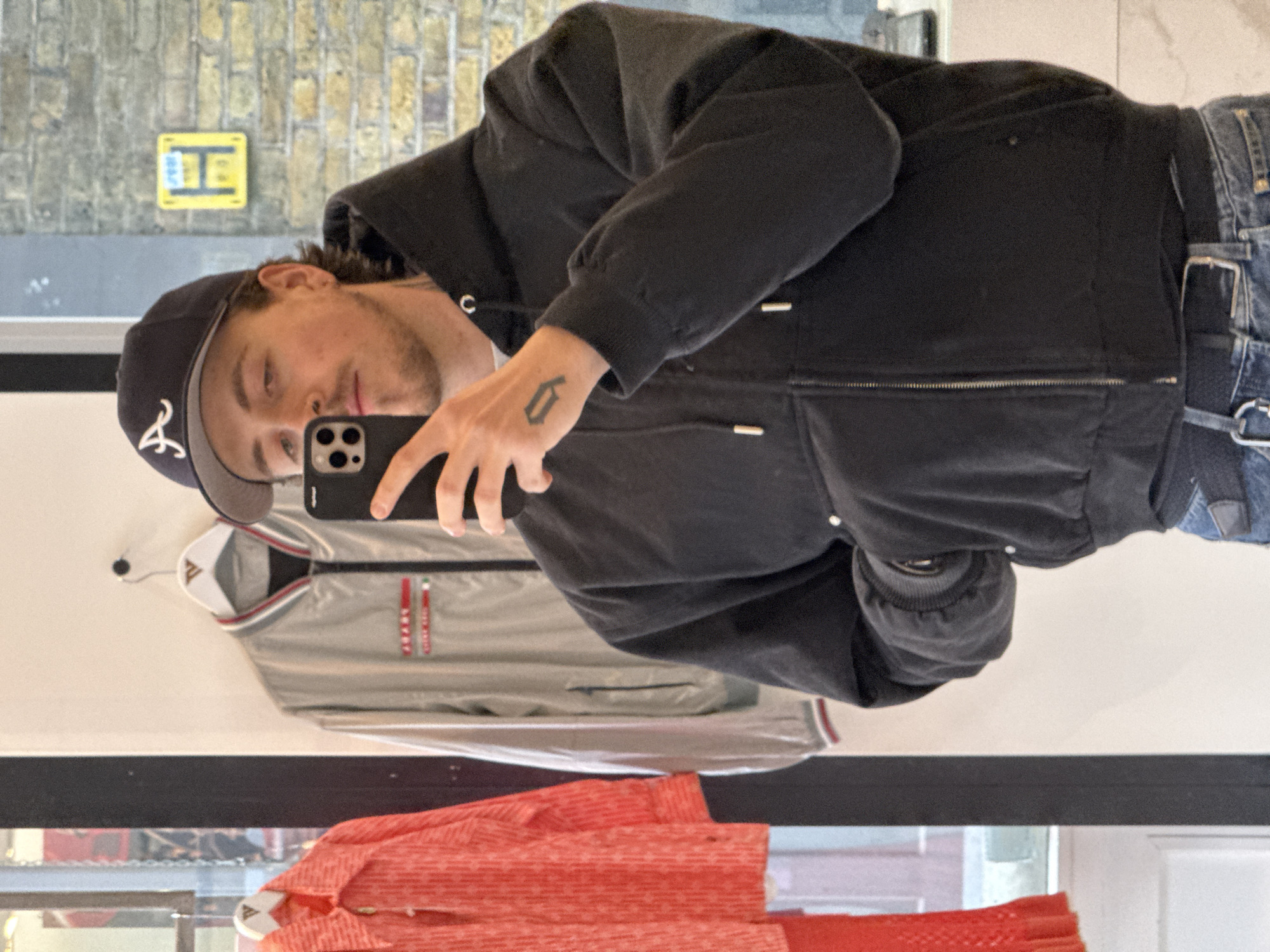 A man is taking a selfie in front of a shop window, presumably in London, United Kingdom, as suggested by the presence of a yellow sign typically used to indicate parking restrictions. The man is wearing a black jacket, blue jeans with a black belt, and a baseball cap with the letter "A" on it. He is holding a phone up to his face with his left hand; a small tattoo is visible on the back of his hand. In the reflection, behind the man, are two items of clothing hanging on hangers within the shop: a grey Prada jacket and an orange shirt or jacket. The environment appears to be an urban setting with a brick wall visible through the reflection in the shop window. The window also reflects the outside environment.
