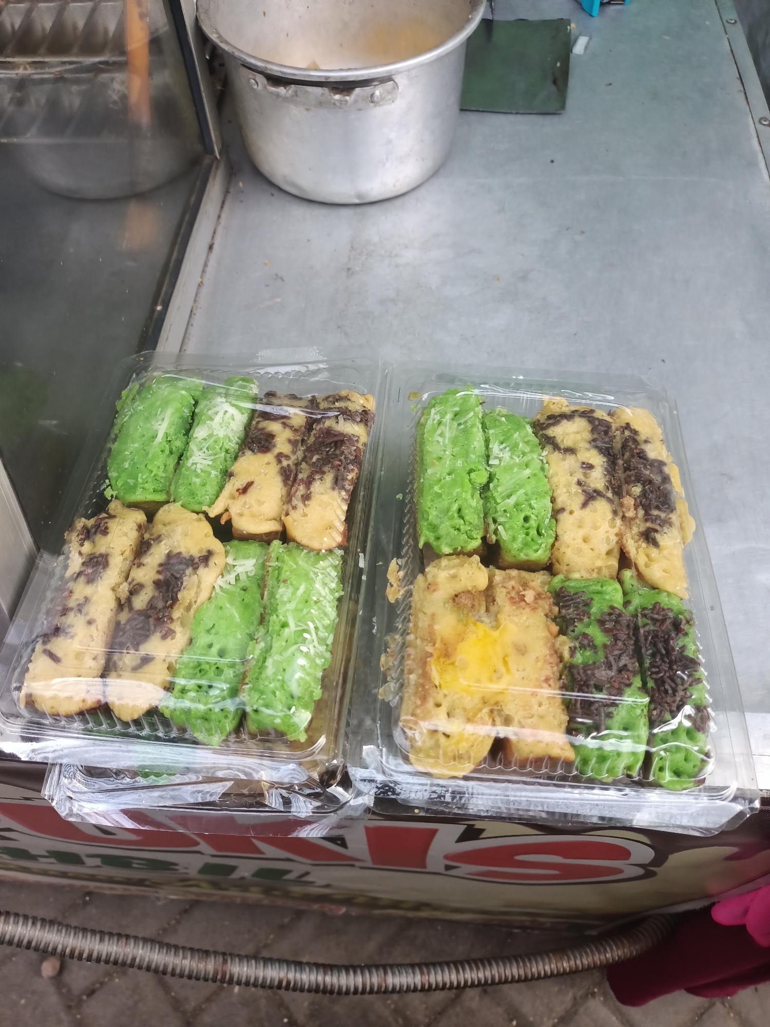 Two clear plastic containers filled with Indonesian snacks, likely Kue Pukis, are displayed on a metal surface. The snacks are arranged in rows and appear to be a mix of green-colored ones, possibly pandan flavored, and yellow-colored ones with chocolate sprinkles and some white grated cheese. Some of the yellow snacks have a more solid, eggy appearance, suggesting a different variation or a filling. The containers are positioned on a cart or food stall, evidenced by the blurry logo at the bottom of the image that contains Indonesian text, possibly indicating it is located in Badas, Indonesia. In the background, a large metal pot is visible, further suggesting a food preparation or serving area. The overall lighting is natural and the scene appears to be an outdoor or semi-outdoor market setting.
