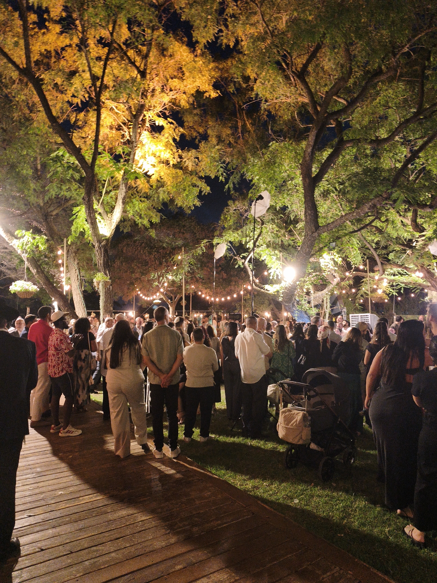 The image depicts an outdoor gathering at night in the city of Hulda, Israel, likely a wedding or reception. A crowd of people, dressed in semi-formal attire, are mingling on a wooden walkway and on the grass. String lights illuminate the trees and the scene, creating a festive atmosphere. The trees are lush and green, with some foliage showing yellow and orange hues, possibly due to lighting or the season. A stroller is visible on the grass, suggesting the presence of families. In the background, more lights and structures can be seen, indicating the event is taking place in a well-decorated outdoor venue. The overall impression is one of a lively and pleasant evening event.