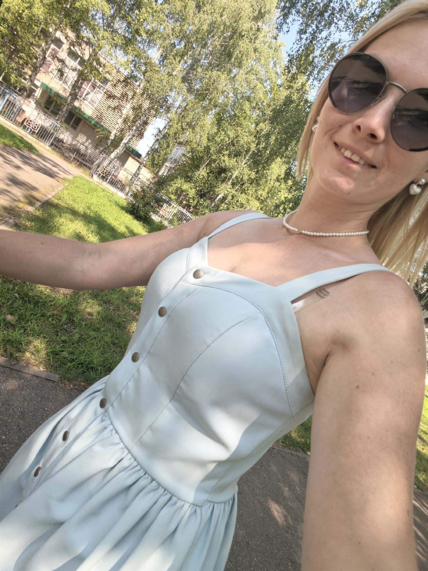 A blonde woman, wearing a light blue dress, pearl necklace, and large sunglasses, smiles at the camera. She is standing outdoors on a sunny day, with trees and a building visible in the background. The setting appears to be a park or residential area in Sterlitamak, Russia, based on the architectural style of the building and the presence of birch trees.