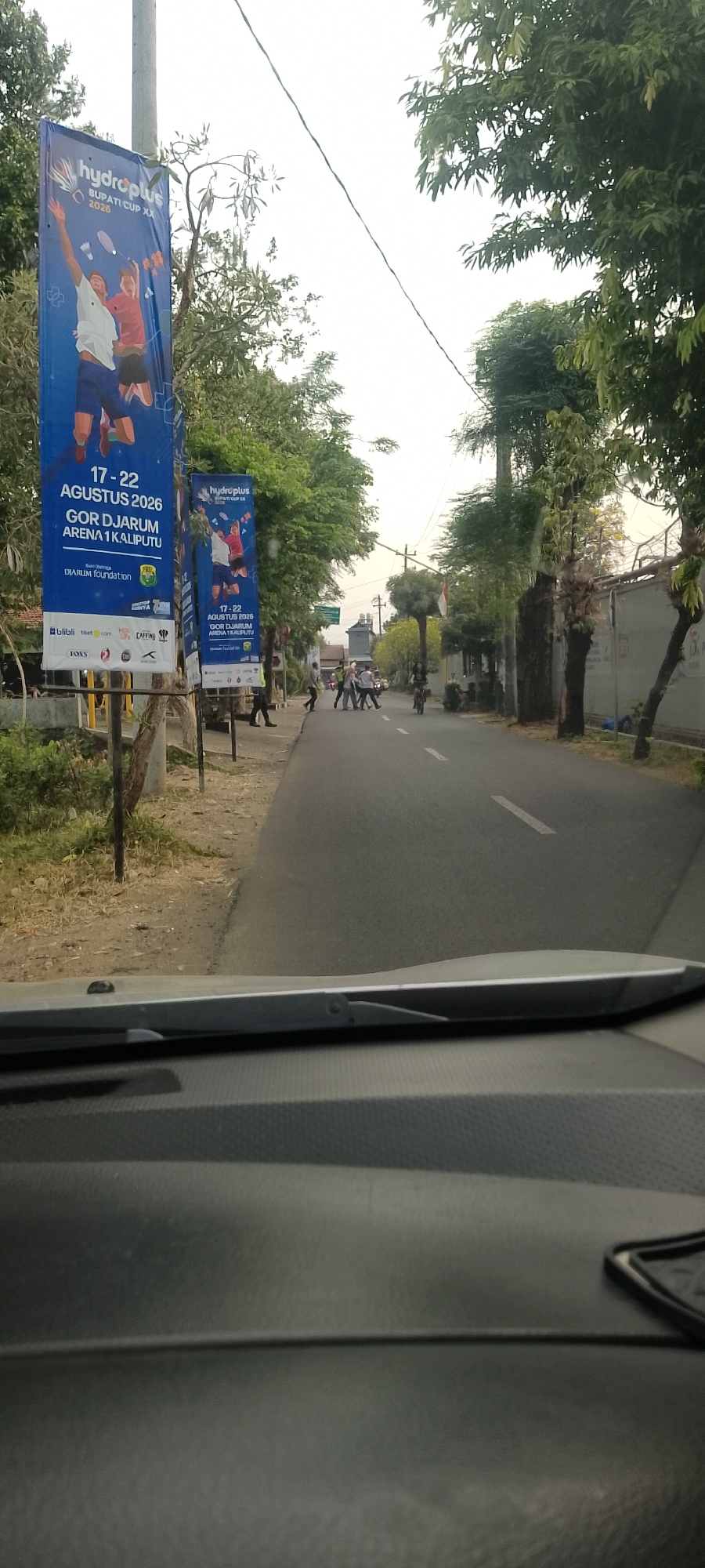 The image shows a street scene in Burikan, Indonesia. In the foreground, a car's dashboard is visible. On the left side of the street, two large blue banners are displayed on poles. These banners advertise the "Hydroplus Bupati Cup XX" badminton tournament, which is scheduled to take place from August 17-22, 2026, at GOR Djarum Arena I, Kaliputuh. The banners feature illustrations of badminton players. Further down the street, several people are crossing the road, and a person is riding a bicycle. The street is lined with trees on both sides, and there are buildings visible in the background. The sky is overcast.