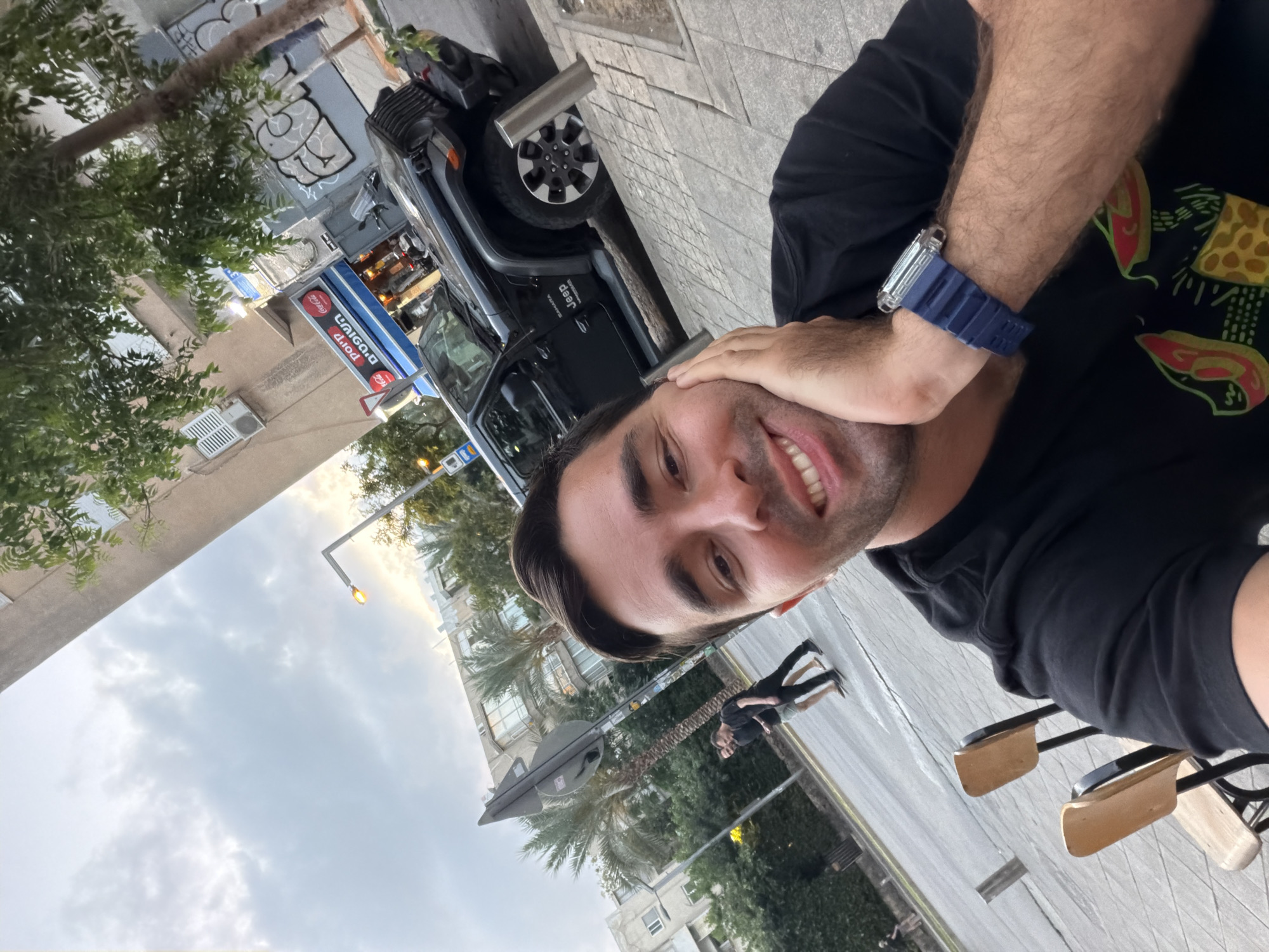 A selfie photograph taken in Tel-Aviv, Israel, shows a man with short dark hair, wearing a black t-shirt and a blue wristwatch. He is smiling and has one hand near his face. Behind him is a black Jeep Wrangler parked on the street. In the background, there is a street scene with buildings, palm trees, and street signs. A couple walks along the sidewalk. There is a Coca-Cola sign visible in Hebrew. The sky is overcast. There are two chairs visible in the foreground. The image is rotated clockwise.