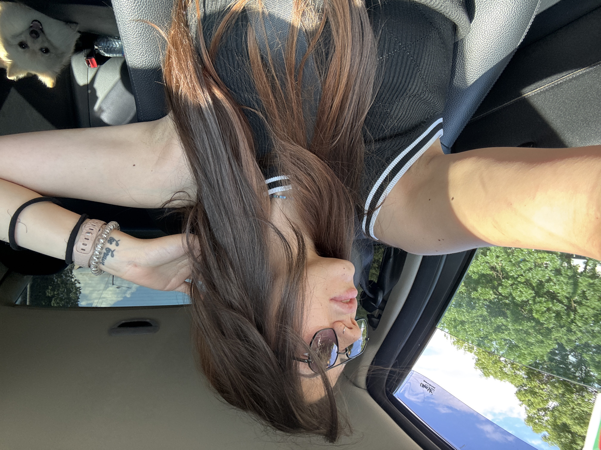 A woman with long brown hair, wearing sunglasses, a black ribbed top with white stripes on the neckline, and a necklace, is in the passenger seat of a car. She is looking out the window to her left, with her arm resting on the open window frame. A small, light-colored dog is visible in the back seat. The scene appears to be a sunny day, with green trees and a blue sky visible through the window. The car interior is dark, with black leather seats. The photo was taken in the city of Sofia, Bulgaria.