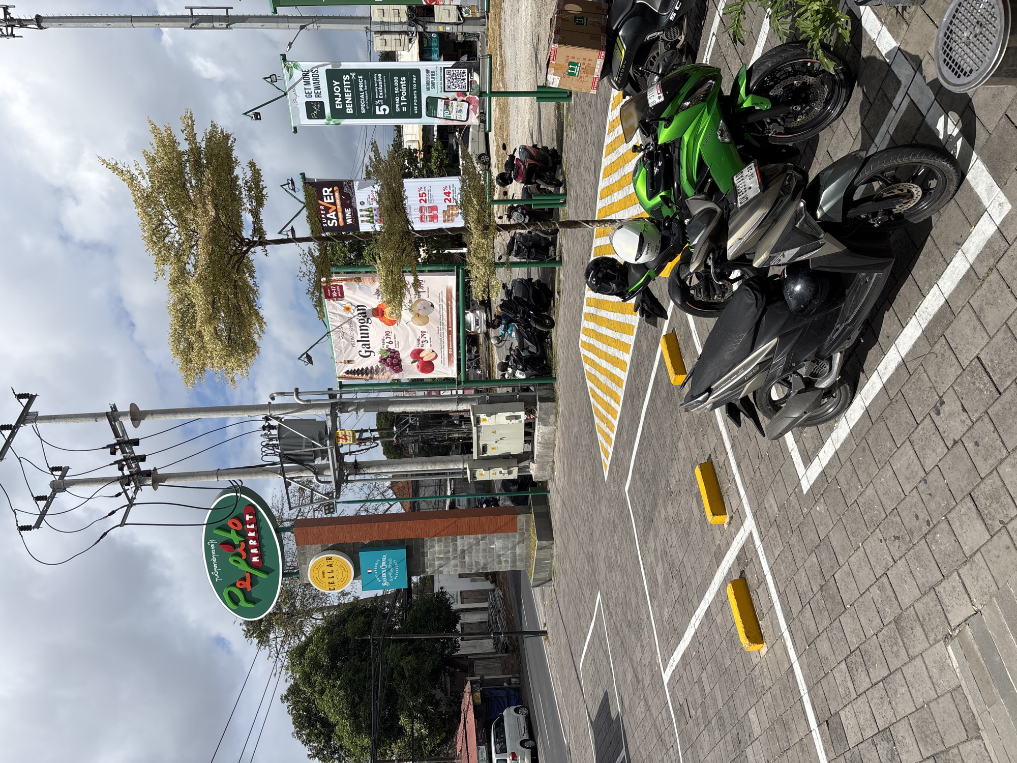 The image depicts an outdoor parking lot in front of a market or commercial establishment in Kuta Selatan, Indonesia. In the foreground, two parked motorcycles are visible: a dark grey scooter in the lower left and a bright green sport motorcycle behind it, partially obscuring the scooter's rear. Both motorcycles are parked within marked parking bays delineated by white lines and yellow parking curbs. The ground surface of the parking lot is paved with textured grey interlocking bricks.
In the mid-ground, several other motorcycles are parked in a row. Behind the parked motorcycles, a street is visible with a white vehicle driving on it. Further back, there are trees, buildings, and signage. A prominent green oval sign with red lettering that reads "Pepito MARKET" is mounted on a brick pillar. Other signs in the background include banners advertising "Galungan," "SUPER SAVER WINE," and "ENJOY BENEFITS." Electrical poles and wires are also prominent elements of the scene, stretching across the upper portion of the image against a cloudy sky. The overall environment appears to be a sunny day with scattered clouds.