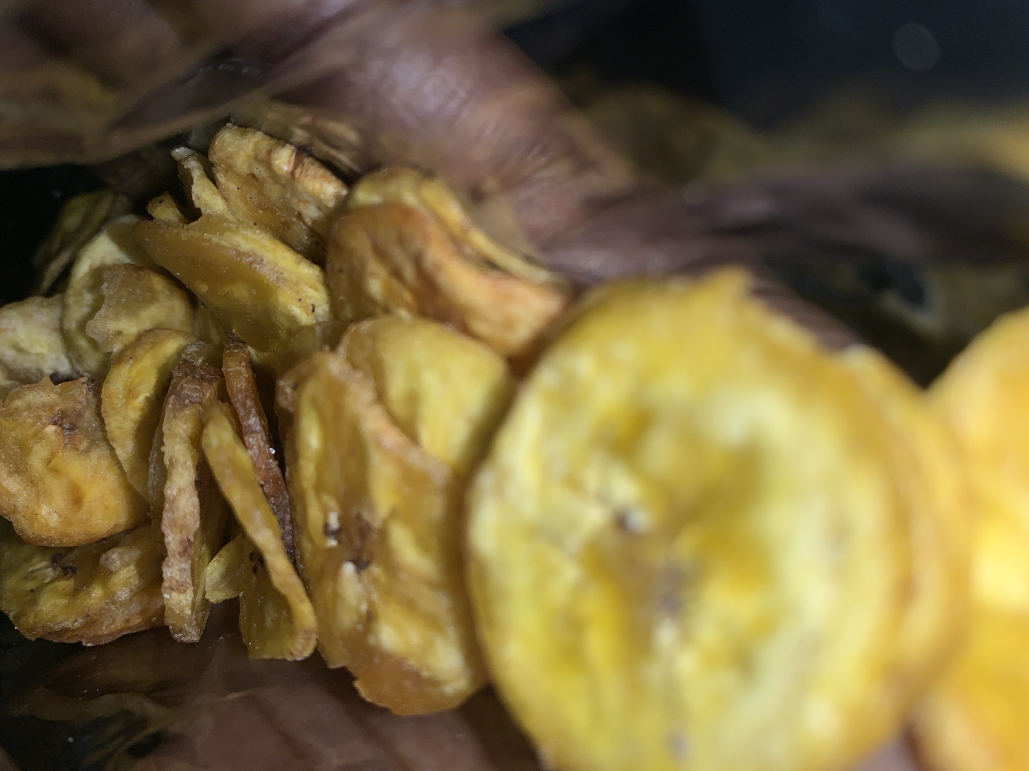 A close-up shot of dried banana chips inside a container or bag. Some of the chips appear darker and crisper than others. The background is blurred, but what appears to be skin is visible at the top of the frame. Location: Unidentifiable, Rafin Par, Nigeria is not confirmed.