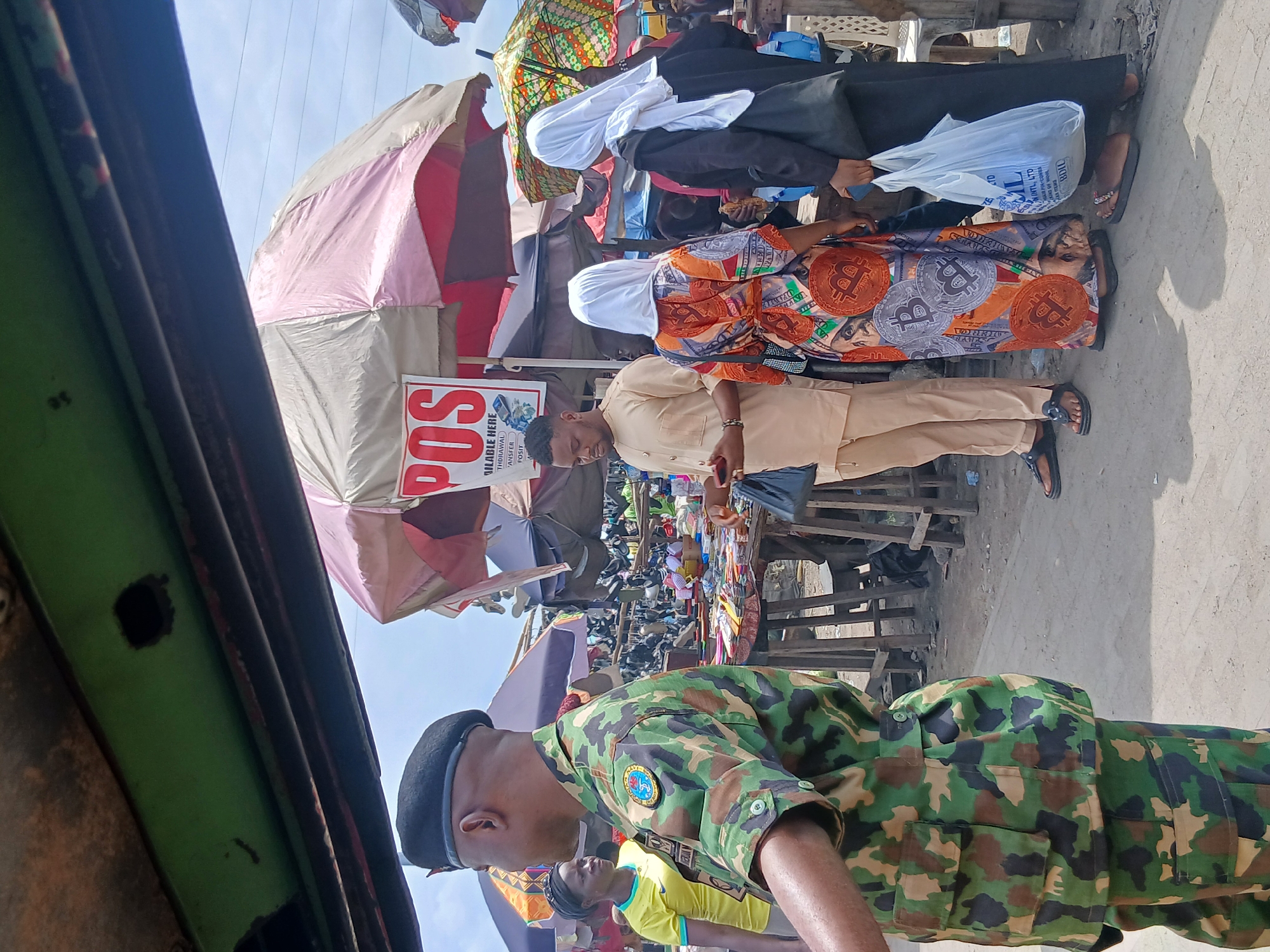 The image depicts an outdoor market scene in Agani, Nigeria. In the foreground, a person in camouflage military uniform is partially visible, facing right. Behind them, a group of people are gathered around a stall with umbrellas. One individual, wearing a tan outfit, holds a mobile device. Two women in headscarves are also present; one wears a colorful dress adorned with Bitcoin logos, and the other holds a white plastic bag. Various goods are displayed on tables and racks in the background, suggesting a busy marketplace. The environment appears to be a dusty, open-air area with stalls and a cloudy sky overhead.
