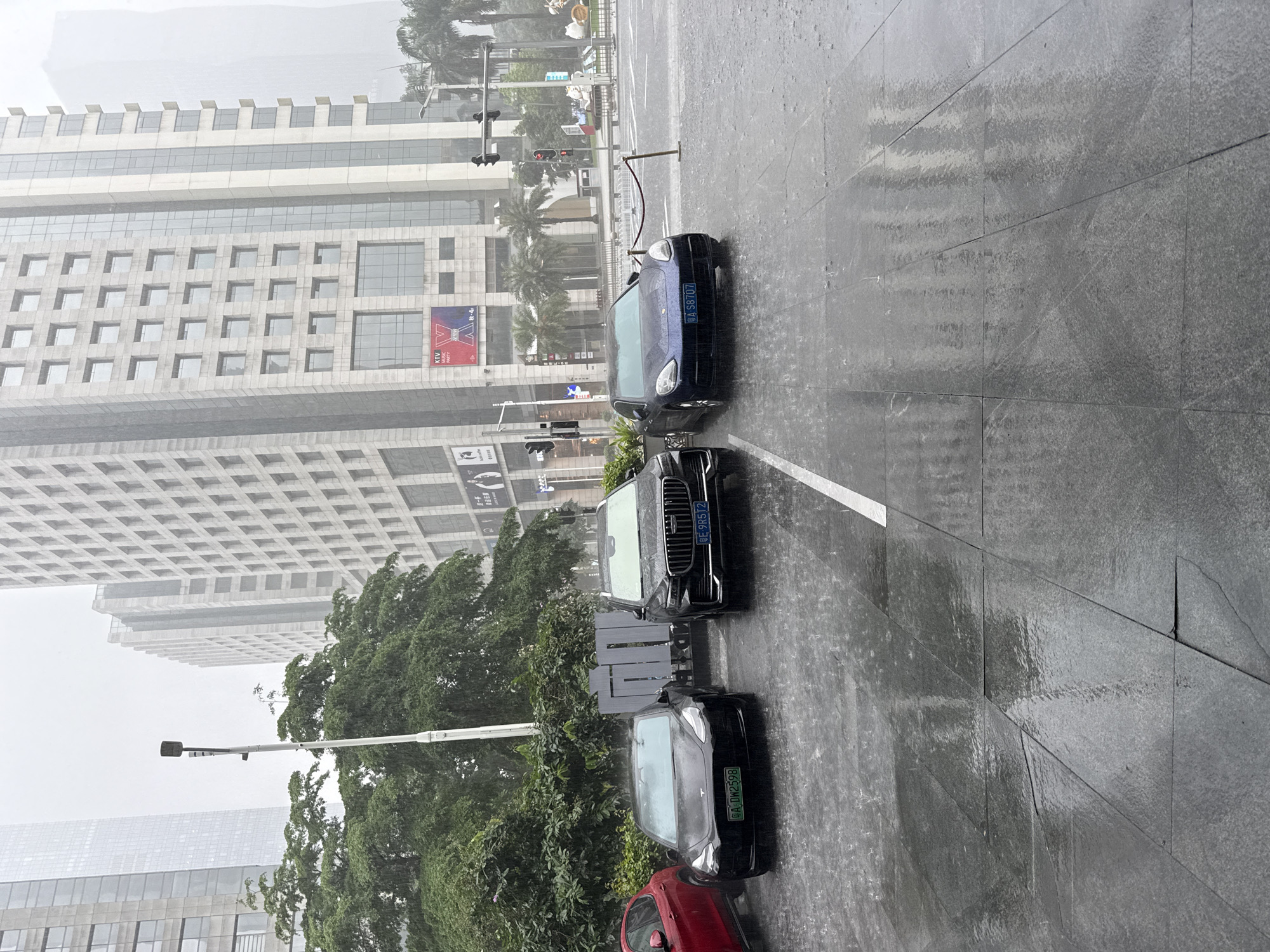 The image depicts a street scene in Tianhe District, China, during a heavy downpour. Several vehicles are parked on the wet asphalt, including a black Tesla, a black Volvo SUV, and a dark blue Porsche. The Tesla has a license plate that reads "A DW2598", and the Volvo's license plate is "CE-9R5T2". The Porsche's license plate is "A S8707". In the background, tall, modern buildings with glass facades rise above lush green trees. Traffic lights and street lamps are visible, and the ground is covered in puddles from the rain, with water droplets clearly visible in the air and on the surfaces. The overall atmosphere is one of a rainy day in a bustling urban environment.