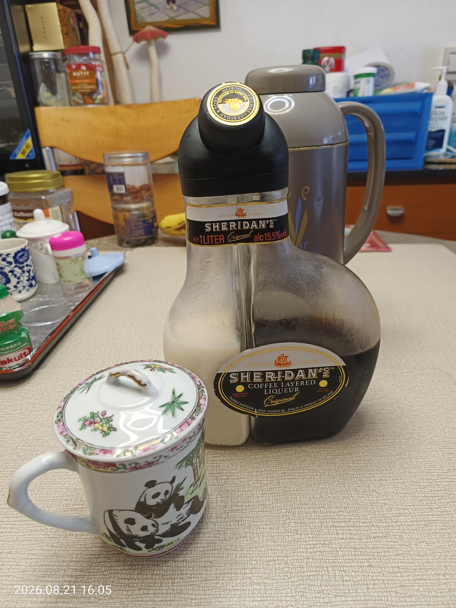 The image shows a table with several items, including a bottle of Sheridan's coffee-layered liqueur, a mug with a panda design and a lid, a thermos, and various jars and containers in the background. The liqueur bottle is distinctive, with two separate compartments for the coffee and cream layers. The mug is white ceramic with a detailed panda illustration and a decorative lid. In the background, there is a wooden chair, a blue plastic container, and some framed pictures on the wall. The overall impression is a domestic setting, possibly a home or an office pantry. The timestamp "2026.08.21 16:05" at the bottom left corner indicates the date and time the image was taken.