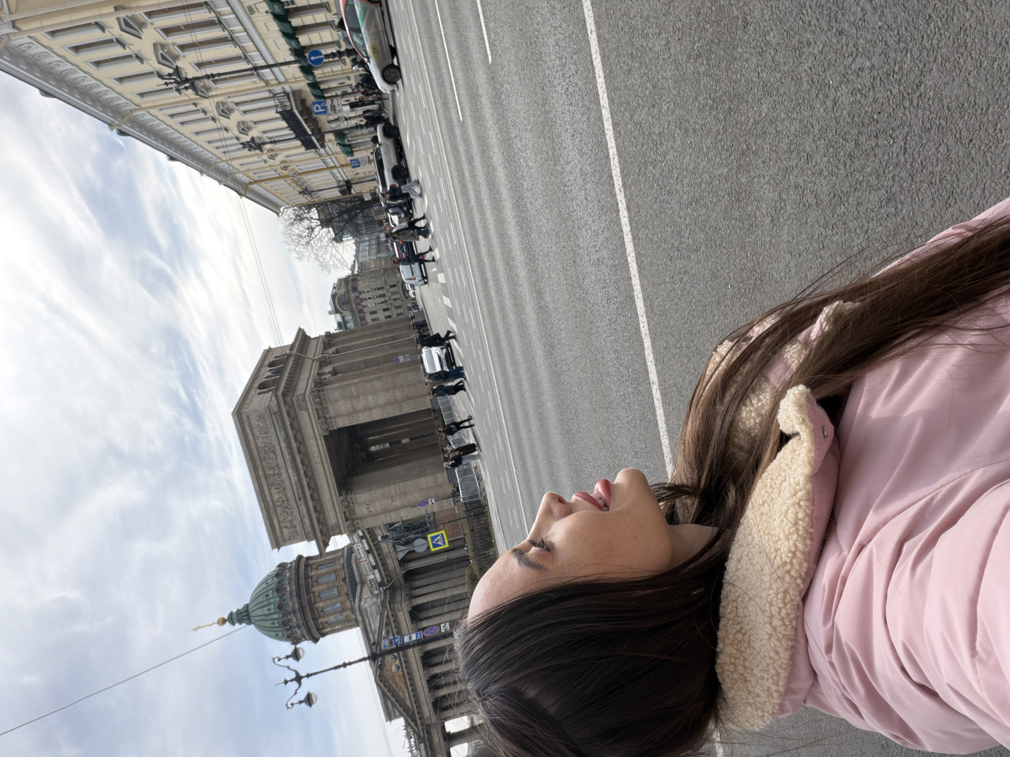 A woman wearing a pink jacket with a white fleece-lined hood is lying on the sidewalk. In the background is the Kazan Cathedral, a Russian Orthodox church in Saint Petersburg, Russia. Beyond that, a city street with cars, pedestrians, and buildings is visible. The sky is overcast with pale clouds.