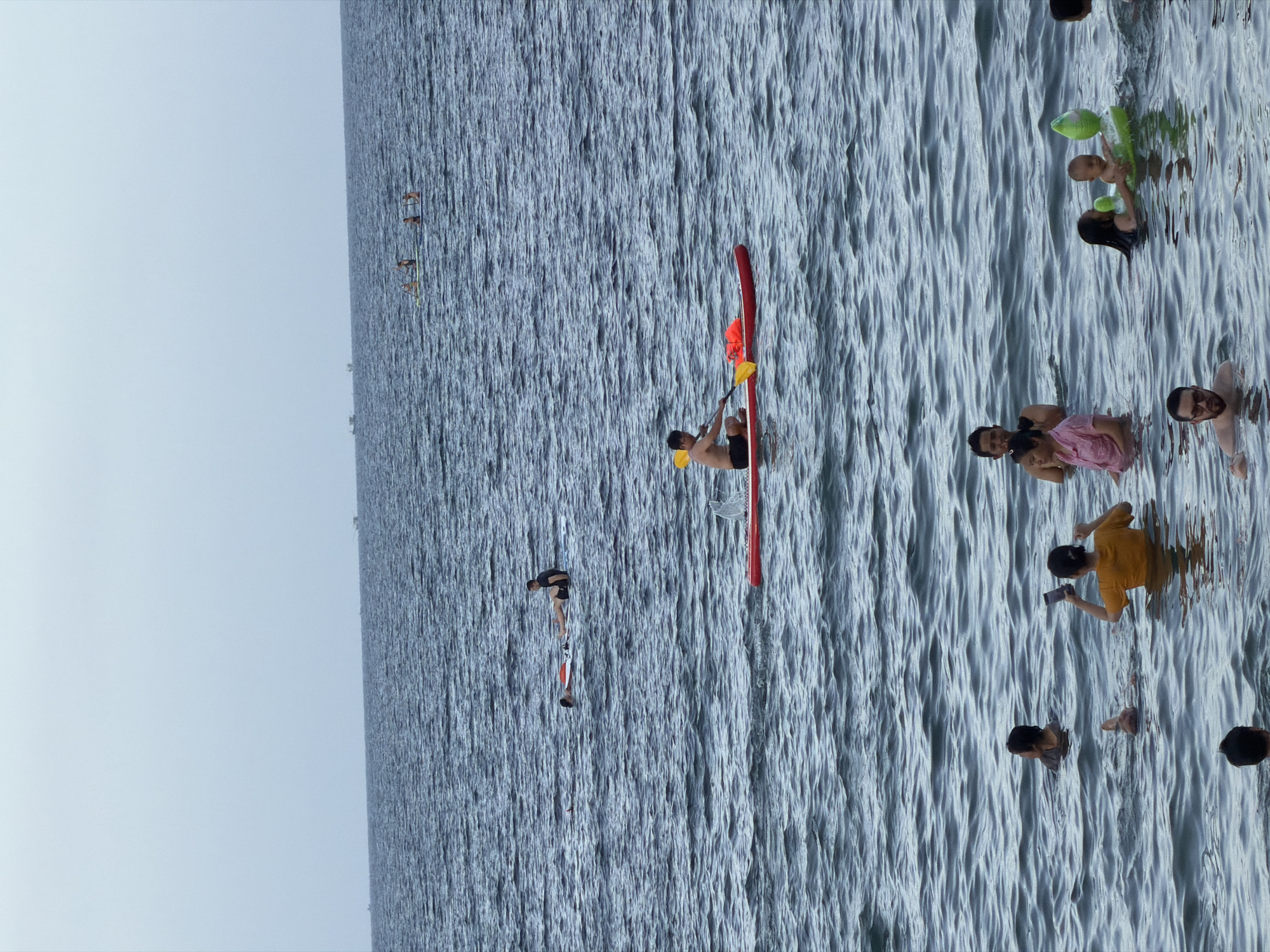 This is a photograph taken on a cloudy day at a beach in Nha Trang, Vietnam. The image shows a group of people enjoying various water activities. In the foreground, several people are wading in the ocean, including a woman in an orange shirt taking a picture with her phone, and two children in a green inflatable toy. A man with glasses is also visible in the foreground, partially submerged in the water. Further out in the water, a man is paddleboarding on a red board, holding a yellow paddle and a life vest. In the middle ground, another person is lying on a paddleboard, with a second person sitting on it. In the background, several other individuals are on paddleboards, and a few distant boats are visible on the horizon. The water is dark blue and choppy, with moderate waves. The sky is overcast with a pale blue-grey hue.