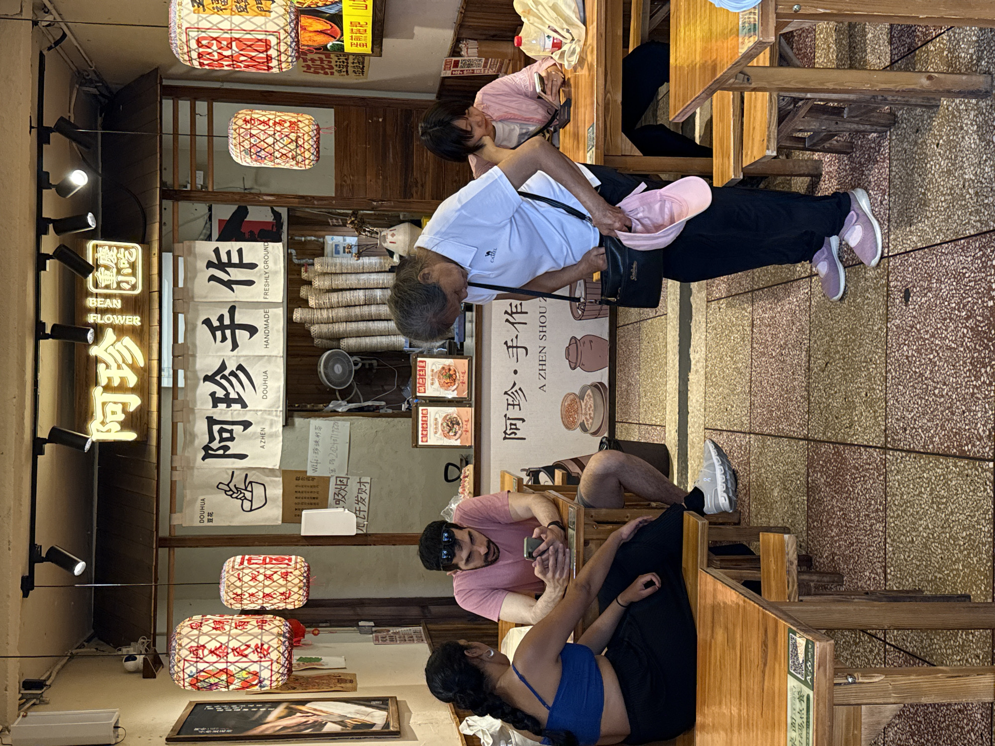 Here is a detailed description of the image:
**General Overview:**
The image shows the interior of a restaurant or cafe, with several people seated at wooden tables and chairs. The overall atmosphere appears casual and the lighting is a mix of natural light and decorative lanterns.
**People:**
* A couple is sitting at one of the tables. The man is wearing a pink shirt, sunglasses, and is looking at his phone. The woman is wearing a blue top and black pants and appears to be looking at something on the table.
* An older woman is standing and seems to be getting something out of her purse. She is wearing a white shirt and black pants.
* Another woman is sitting at the table in the background, wearing a pink shirt.
**Objects and Environment:**
* **Restaurant Details:** The restaurant has a rustic, traditional Chinese theme. The tables and chairs are made of wood. The floor is tiled with a mosaic pattern.
* **Signage:** There are several signs in both Chinese and English, including "Bean Flower" and "Azhen Shou Zu". There is also a poster with images of food.
* **Decorations:** There are decorative lanterns hanging from the ceiling. Also hanging are banners written in Chinese.
* **Fans:** There are several decorative fans on display.
**Actions and Interactions:**
* The couple at the table seems to be engaged in their own conversation or activities, possibly looking at their phones or talking.
* The older woman is in the process of retrieving something from her bag.
**Location:**
Based on the signage in Chinese characters and the overall aesthetic, the location is likely in Yuzhong District, China.