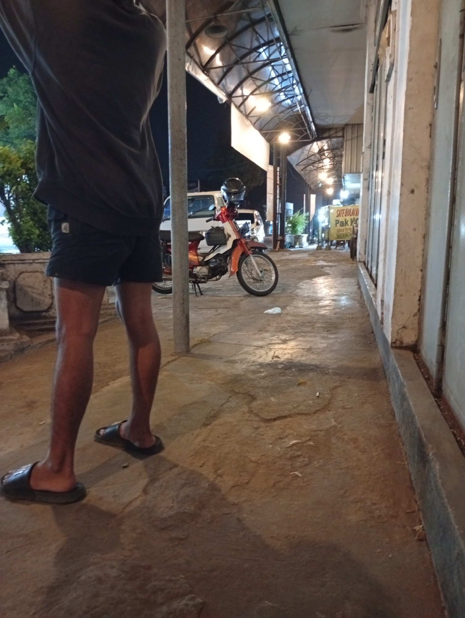 A person, whose lower body is visible from the knees down, stands on a concrete sidewalk at night. They are wearing black shorts and black flip-flops. The person is facing away from the camera and towards a street. To their right, a motorcycle is parked on the sidewalk. The motorcycle is orange and white with a black seat and a black basket on the front. Behind the motorcycle, a white van is visible. Further down the street, a yellow sign with red lettering is mounted on a pole. The sign has text that is difficult to read but appears to be advertising food. The sidewalk is illuminated by overhead lights, which cast shadows on the ground. Trees and foliage are visible in the background, to the left of the frame. The overall scene suggests an outdoor setting at a commercial establishment or street vendor.