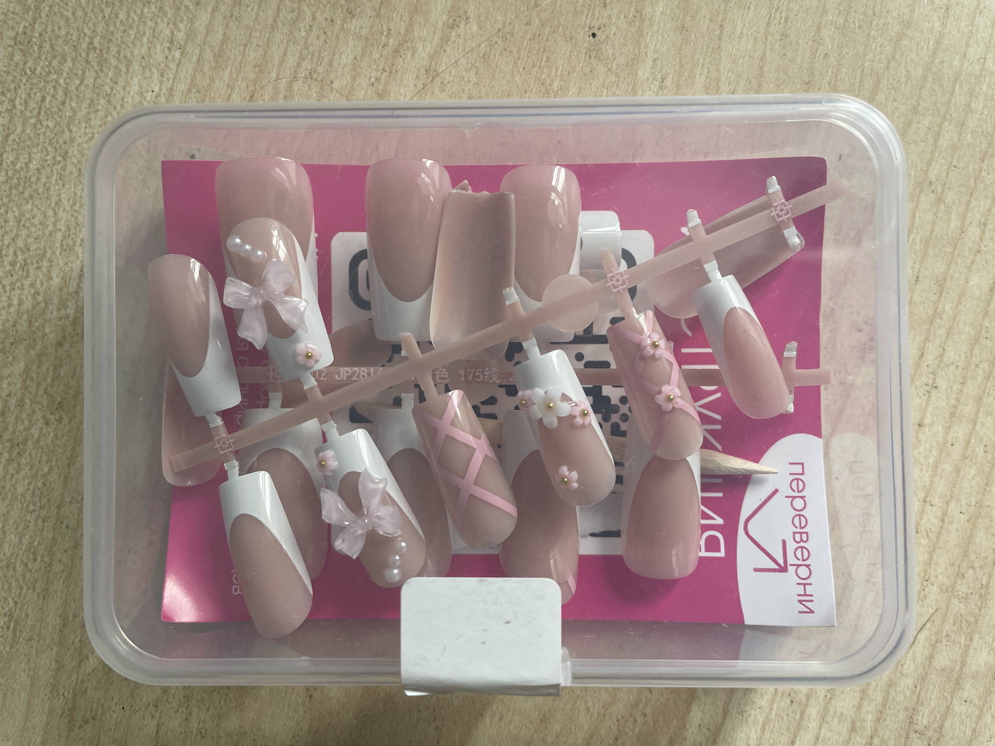 A set of artificial nails, many adorned with bows, pearls, and floral accents, are neatly arranged in a clear plastic container on a light brown wooden surface. The container is positioned on a pink insert card with some text visible, including what appears to be Russian writing. The nails are in various shades of pink and white, with some featuring French tip designs and others decorated with intricate pink ribbon patterns. The overall scene suggests a retail or personal collection setting. The provided crops offer close-up views of the nail details and container. There is no information within the image to indicate a specific location in Vologda, Russia.