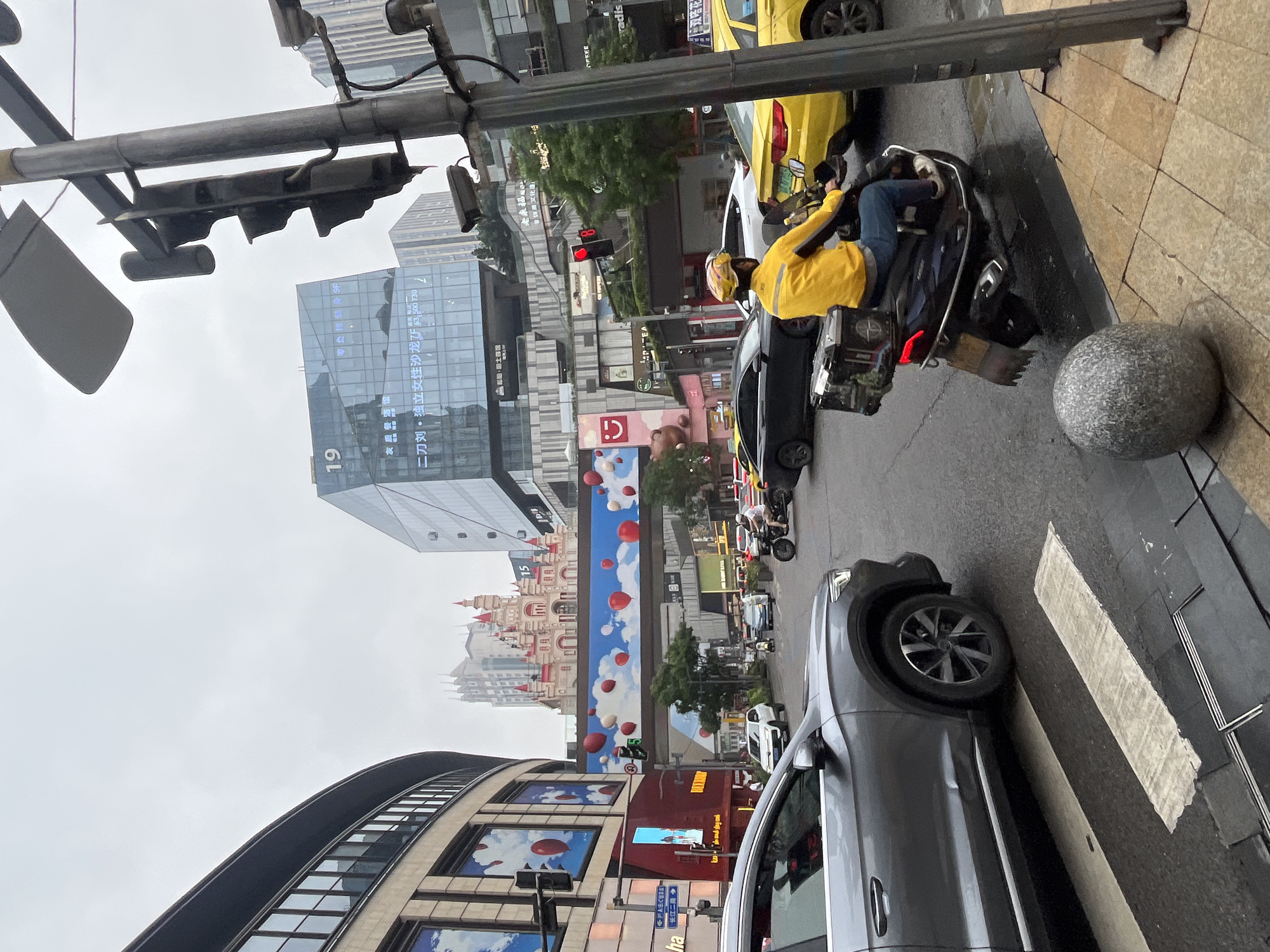 The image depicts a street scene in Yuzhong District, China, likely on a cloudy day given the overcast sky. In the foreground, a gray SUV is stopped at a crosswalk. To the right, a person wearing a bright yellow jacket and blue pants rides a scooter with a black cargo box on the back. They are in motion, heading towards the right side of the frame. Further in the background, a yellow taxi is visible, and other vehicles are present on the wet road.
The urban environment is characterized by multi-story buildings with modern architecture, including one with the number "19" displayed prominently. A large, colorful advertisement featuring a cartoon castle and red balloons spans across an overhead walkway. Traffic lights are visible, with one showing a red signal. The sidewalk is paved with square tiles, and a large, gray, egg-shaped bollard is situated on the edge of the walkway. The overall impression is of a busy urban intersection.