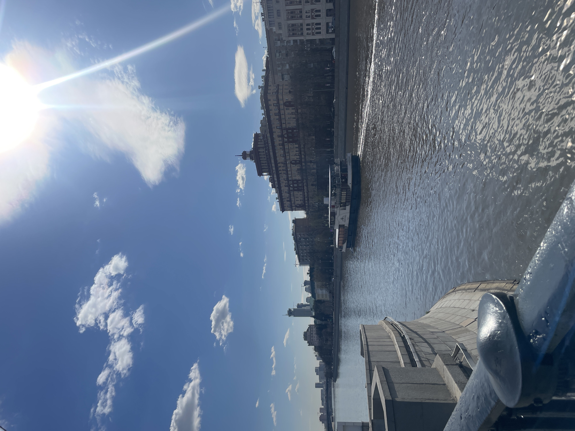The image shows a section of the Moskva River in Moscow, Russia, from an elevated position, likely a bridge. The river flows from top to bottom in the image. A passenger boat is traveling on the river. The buildings along the riverbank suggest an urban environment. The sky is blue with scattered white clouds and a visible sun glare. A section of a bridge railing and stone structure is visible in the foreground on the right side of the image.