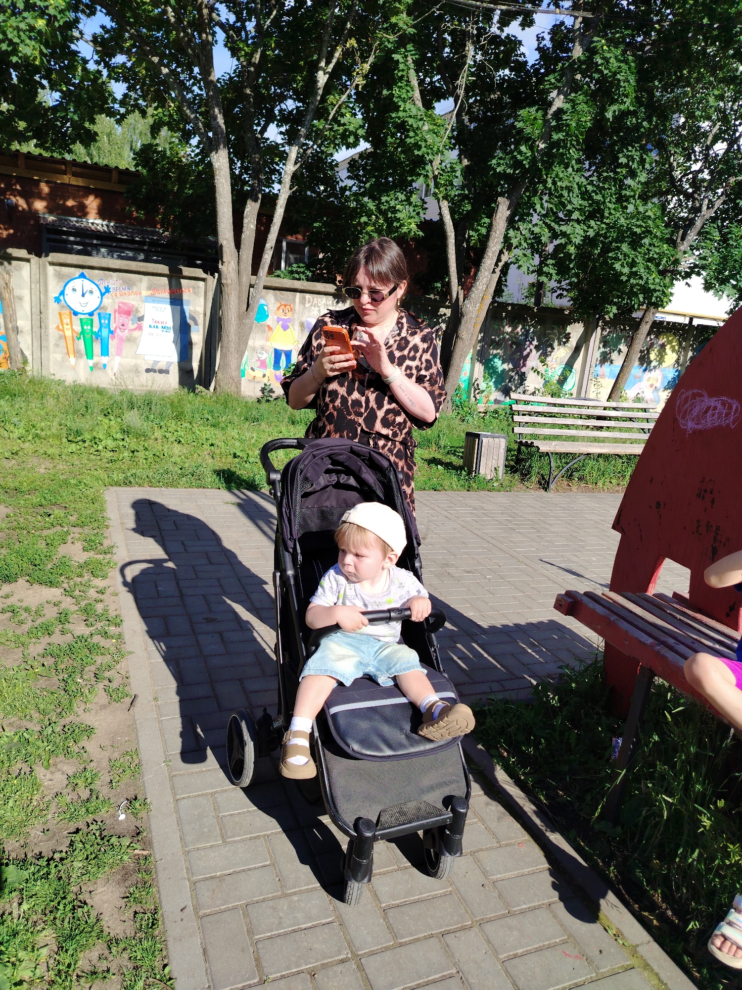 In Noviy, Russia, on a sunny day, a woman wearing sunglasses and a leopard-print top holds a mobile phone. Beside her, a child in a stroller, wearing a white cap and denim shorts, looks to the side. The stroller is on a paved walkway lined with grass and trees. A concrete wall with colorful murals is in the background, along with a wooden bench and a red structure. The overall setting appears to be a park or playground.