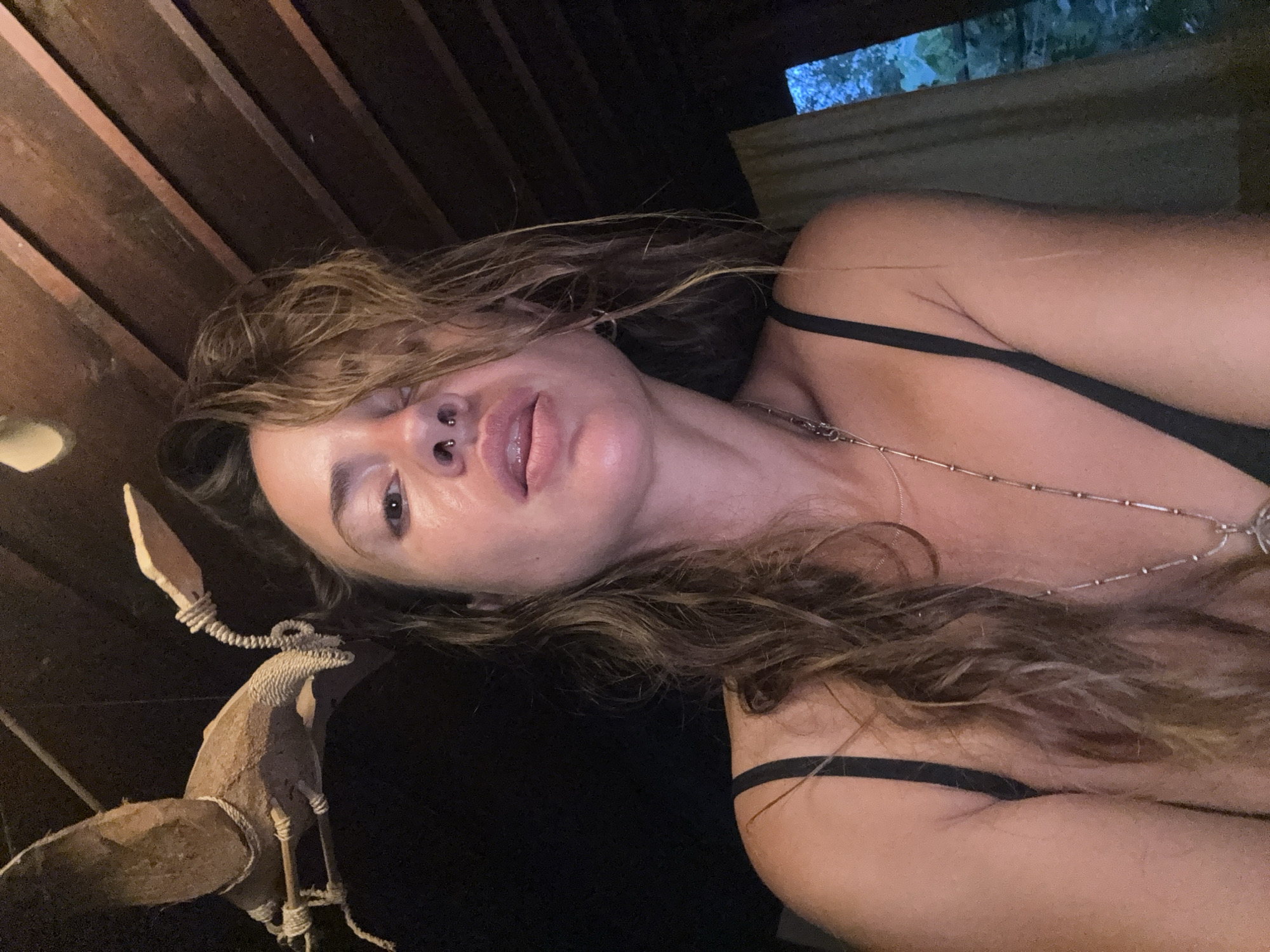 A low-angle, close-up selfie shows a woman with long, wavy brown hair and a nose ring. She is wearing a black bra and a silver necklace. Her head is tilted slightly, and her eyes are looking upwards. Behind her, the ceiling is made of dark wooden planks, and a wooden decoration hangs from the ceiling on the left. To the right, a light-colored curtain is partially visible, revealing a glimpse of green foliage outside. The lighting is dim, with a warm glow on her face. The overall impression is intimate and naturalistic.