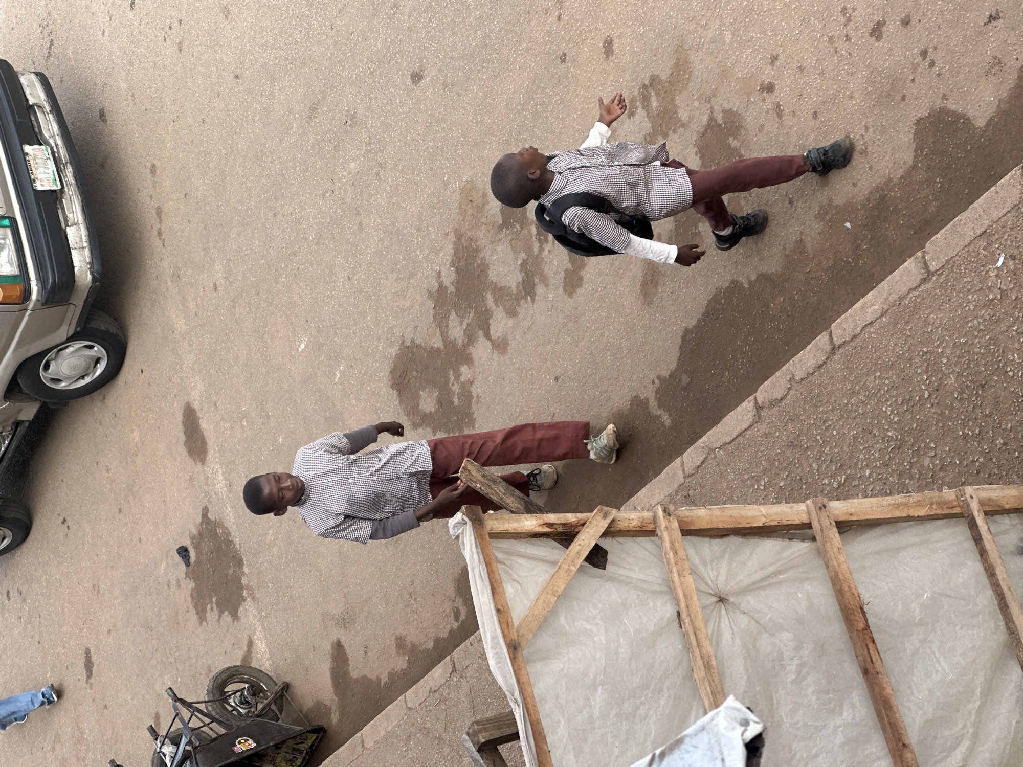 In Bukuru, Nigeria, two young boys walk on a dirt road. The boy on the left wears a checkered shirt, long maroon pants, and green sneakers. He carries a wooden plank in his right hand. The boy on the right wears a similar checkered shirt with a white long-sleeved undershirt, maroon pants, and black sneakers. He has a black backpack on his shoulders and is walking with his right arm extended. They are walking on a paved road that appears to be a street. A parked vehicle, possibly a van, is visible in the background, and a cart with two wheels is on the left side of the road. A makeshift wooden structure covered with white plastic sheeting is positioned along the edge of the road. The road surface shows signs of wetness and some debris. The scene captures a moment of everyday life in a Nigerian town.