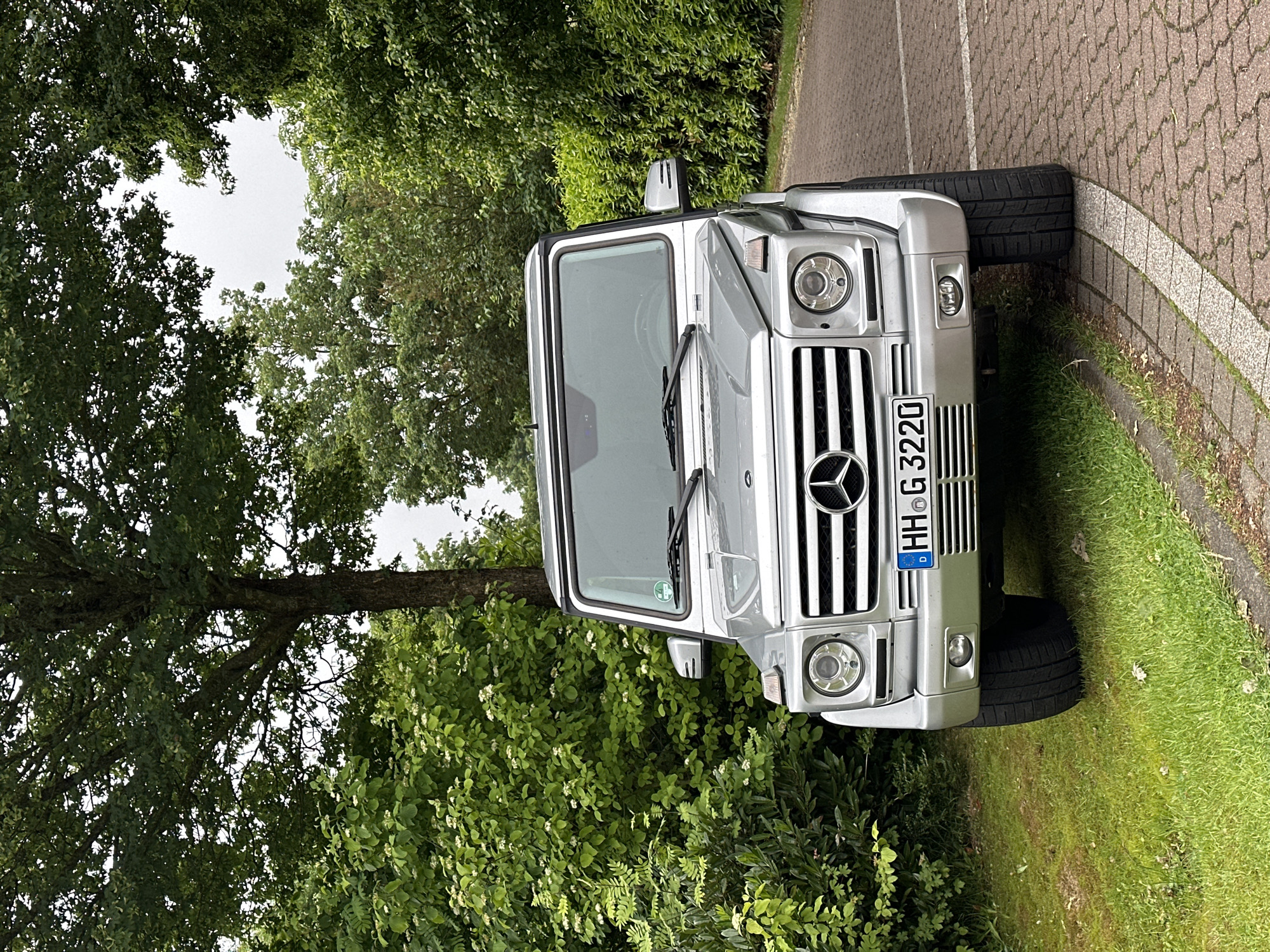 A silver Mercedes-Benz G-Wagen, bearing the German license plate "HH G 3220," is stationary outdoors in Deinste, Germany. The vehicle is positioned facing the viewer, with its front right tire on a reddish-brown brick-paved path and its front left tire on green grass. A prominent Mercedes-Benz logo is visible on its grille, flanked by round headlights. A green environmental sticker is affixed to the passenger side of the windshield. The background features dense green foliage, including a large tree with a thick trunk and a canopy of green leaves directly behind the vehicle. Various bushes and shrubs, some with small white blossoms, surround the car on the left. A neat hedge line borders the paved path on the right side of the image. The sky is overcast, appearing white through gaps in the tree canopy. No individuals are present in the scene.