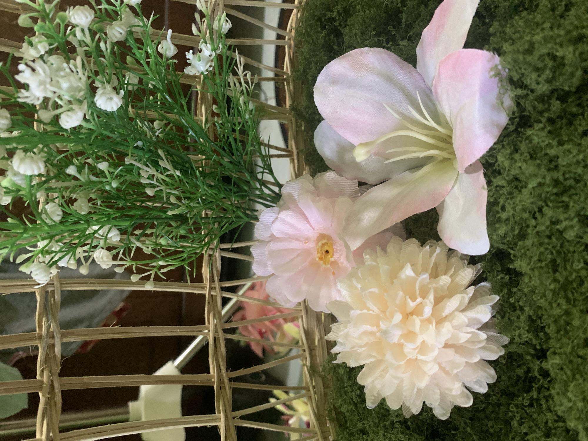 The image shows a close-up view of artificial flowers arranged in a woven basket, placed on a green moss-like surface. In the background, through the basket's weave, out-of-focus pink and yellow flowers are visible. A cluster of small white artificial flowers and greenery is also present, emerging from the basket. The overall scene is still and decorative. There is no indication of a specific location within Kuala Pembuang, Indonesia.