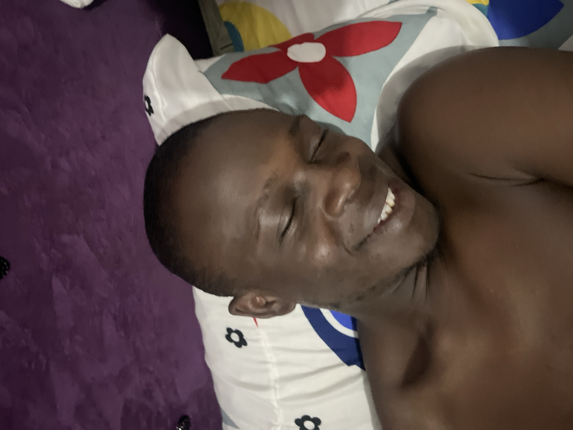 A dark-skinned man is lying on a bed with a pillow. The background includes a purple surface, possibly a blanket or bedspread. A pillow with a flower pattern is under the man's head. The location is implied to be Abuja, Nigeria but is not confirmed by the image alone.