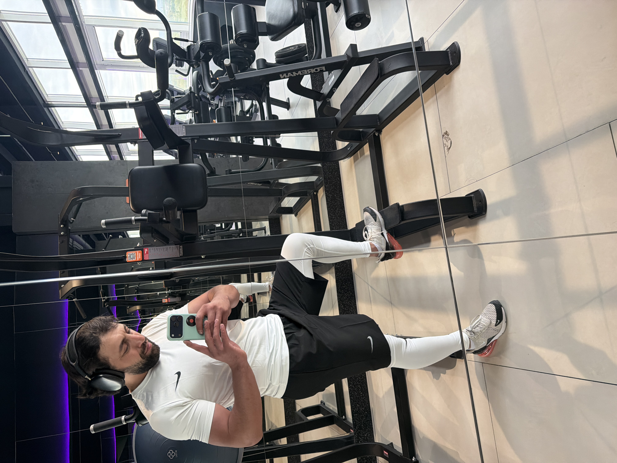 A man is taking a selfie in a gym. He is wearing white leggings, black shorts with a Nike logo, a white t-shirt also with a Nike logo, and headphones. He is sitting on a blue exercise ball and holding a smartphone. In the background is gym equipment, including a Foreman machine. The gym has a mirrored wall, reflecting the scene. The location may be in Krasnoyarsk, Russia.