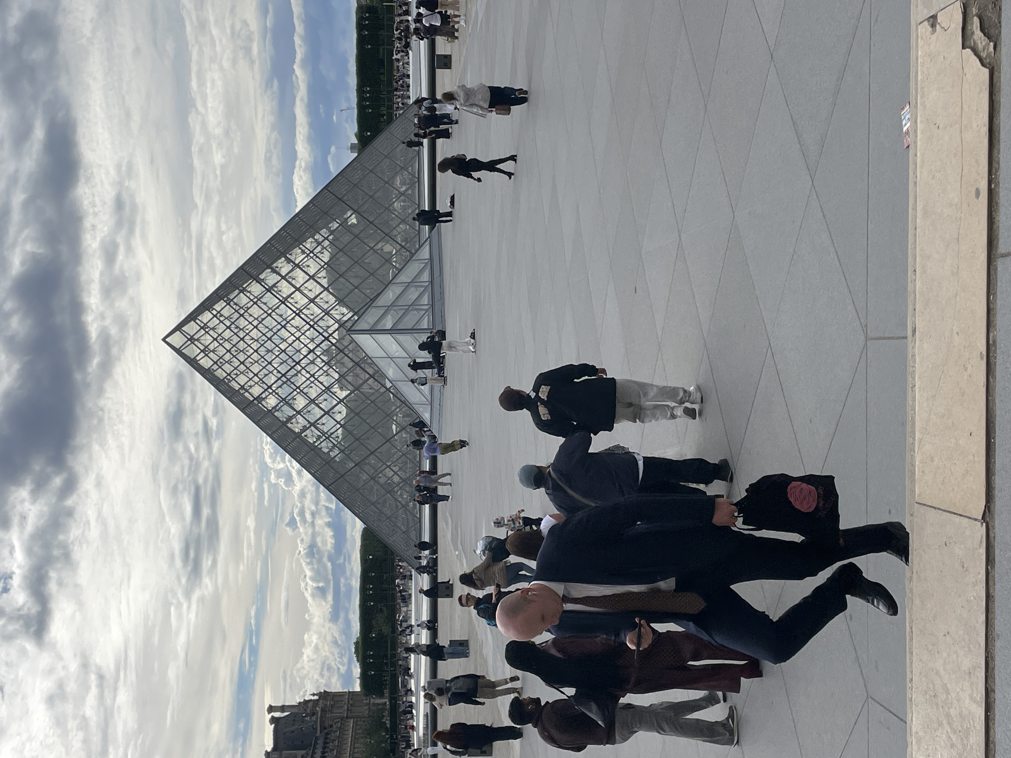 The image depicts the exterior of the Louvre Museum in Paris, France, with the iconic glass pyramid prominently featured in the background. The sky is overcast with dramatic clouds. In the foreground, a man in a dark suit, white shirt, and tie is walking down a set of stairs. He is holding a smartphone in his left hand and carrying a black tote bag with a rose design in his right hand. Several other individuals are visible in the middle ground, walking across the paved plaza in front of the pyramid. Some are dressed casually, while others appear to be tourists. The plaza is large and open, with geometric pavement patterns. In the distance, the historic architecture of the Louvre palace can be seen to the left, and more people are gathered around the pyramid.