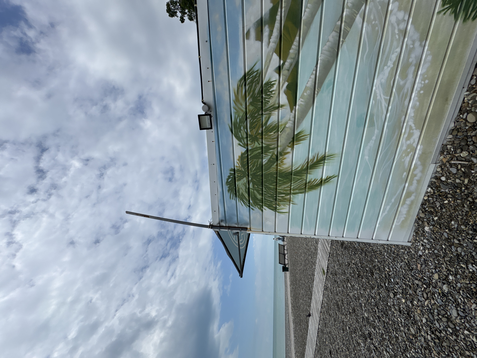 The image shows a beach scene, possibly in Sochi, Russia, based on the presence of a pebble beach and the architectural style of the structure. The sky is overcast with scattered clouds. The structure features a corrugated metal awning painted with a palm tree motif. An angular sign is attached to the awning. A gravel path runs along the edge of the beach. The sea is visible in the distance.