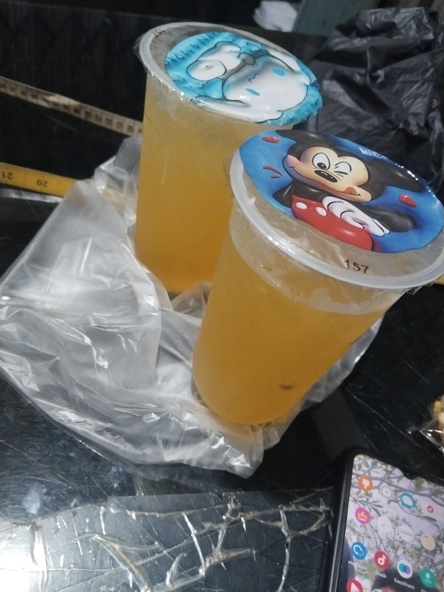 The image depicts two plastic cups filled with yellow liquid, likely a beverage. The cup in the foreground is topped with a lid featuring a cartoon image of Mickey Mouse, with the number "157" printed on the lid's edge. The second cup, positioned behind the first, has a lid with a blue cartoon design, possibly a character from Sanrio. Both cups are partially obscured by clear plastic packaging. A yellow measuring tape with visible markings is present in the background. The surface beneath the cups appears to be dark and textured, possibly a table. A portion of a smartphone with an active screen displaying app icons is visible in the bottom right corner. The setting is indoors, with the background showing a dark, possibly crumpled material. The image quality suggests it was taken with a mobile device in low-light conditions.