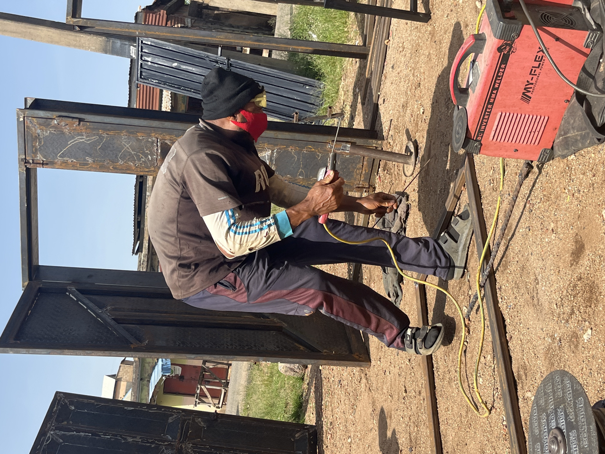 A man wearing a black beanie, red face mask, and a brown jacket is welding metal frames outdoors. He is bent over, holding a welding torch in his right hand, with a bright spark emanating from where it meets a metal rod. He is wearing dark pants with maroon stripes and sandals. The welding equipment, a red DC inverter MMA welder, is visible to the right of the frame, connected to the welding torch by a yellow cable. The ground is covered in reddish-brown dirt and gravel, with various metal bars and a metal cutting disc on the ground. In the background, there are several large, unfinished metal door frames standing upright. Further behind, buildings with corrugated iron roofs and some vegetation are visible. The scene appears to be in an open, urban setting. This image is from Jos, Nigeria.