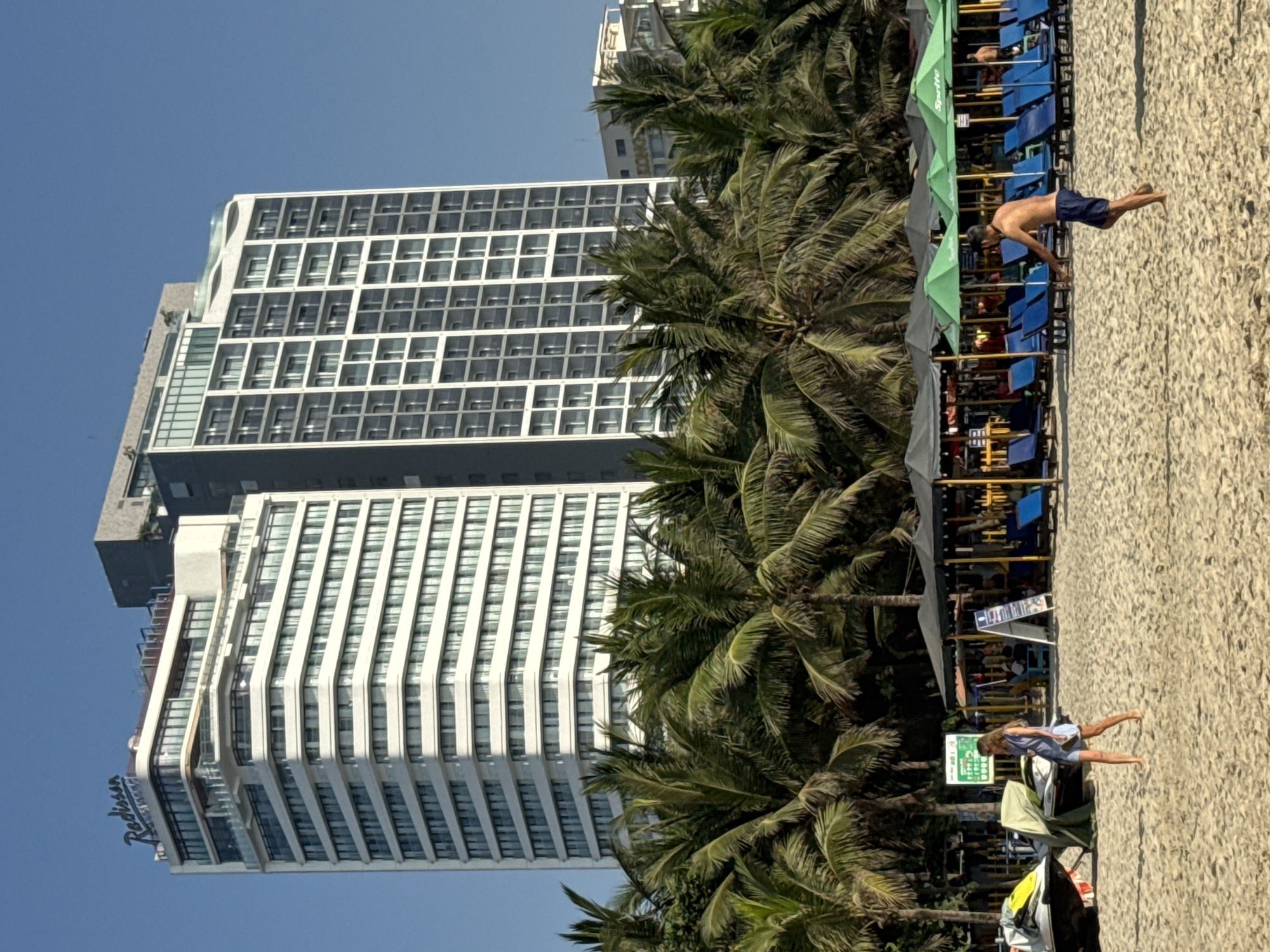 The image depicts a coastal scene in Đà Nẵng, Vietnam, dominated by a tall, modern hotel tower. The hotel, potentially named "Radisson," is partially obscured by palm trees. A beach area extends along the right side of the frame. Two people are visible: one walking along the beach, and another running. Beach chairs and umbrellas line the beach. The sky is clear and blue.
