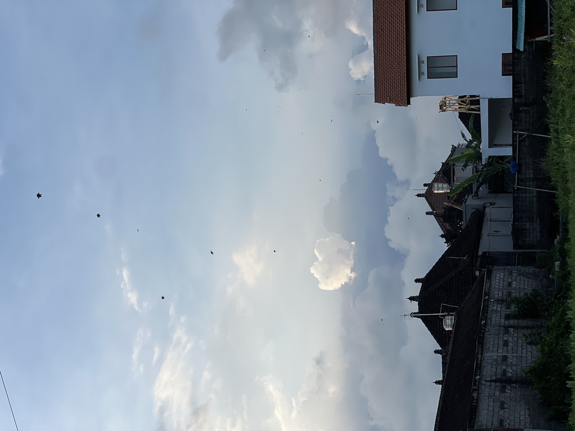 The image captures a daytime scene in Sukawati, Indonesia. The sky is filled with a mix of blue sky and puffy clouds. Several small, dark objects, possibly birds or kites, are scattered across the sky. In the foreground, there are buildings, including a white two-story structure with a red-tiled roof and a traditional Balinese-style building with dark tiled roofs. Green vegetation lines the right side of the frame.