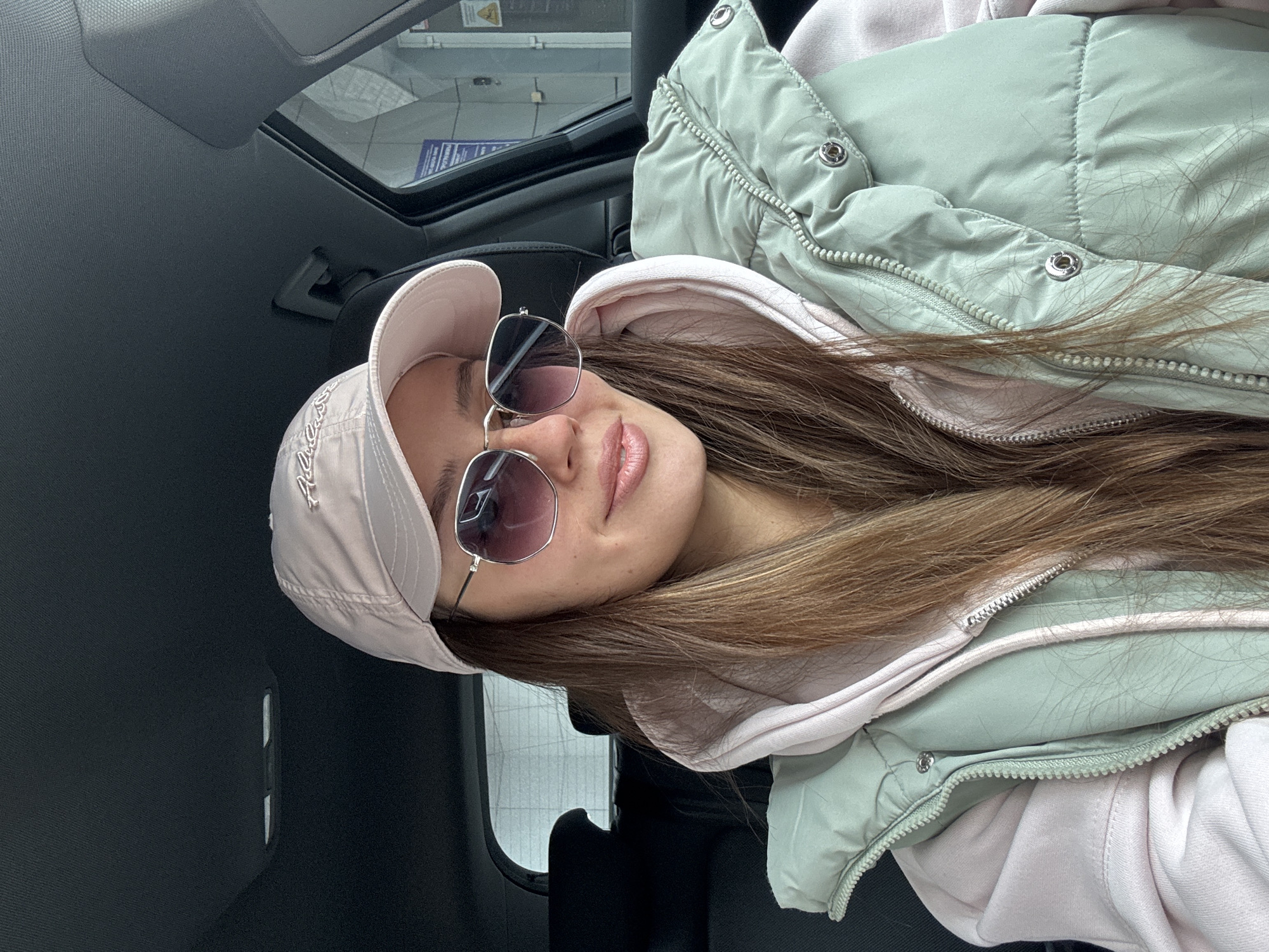 A female individual is depicted seated within a vehicle, appearing to occupy a front passenger or driver's seat. She wears a light pink baseball cap with an embroidered logo, hexagonal sunglasses featuring silver frames and gradient purple lenses, a light pink hooded sweatshirt, and a light mint-green quilted vest with a silver zipper and snap buttons. Her long, brown hair is visible, and she has a slight smile, with pink lipstick applied. The vehicle's interior features dark upholstery. Outside the vehicle's window, a contemporary building is visible. Mounted on this building are multiple signs, including a prominent blue sign with white Cyrillic text, partially legible as "ОТДЕЛЕНИЕ БАНКА" (Bank Branch) and "КРАСНОЯРСК", confirming the location as a bank branch in Krasnoyarsk, Russia. A yellow triangular warning sign is also present on the building's exterior.