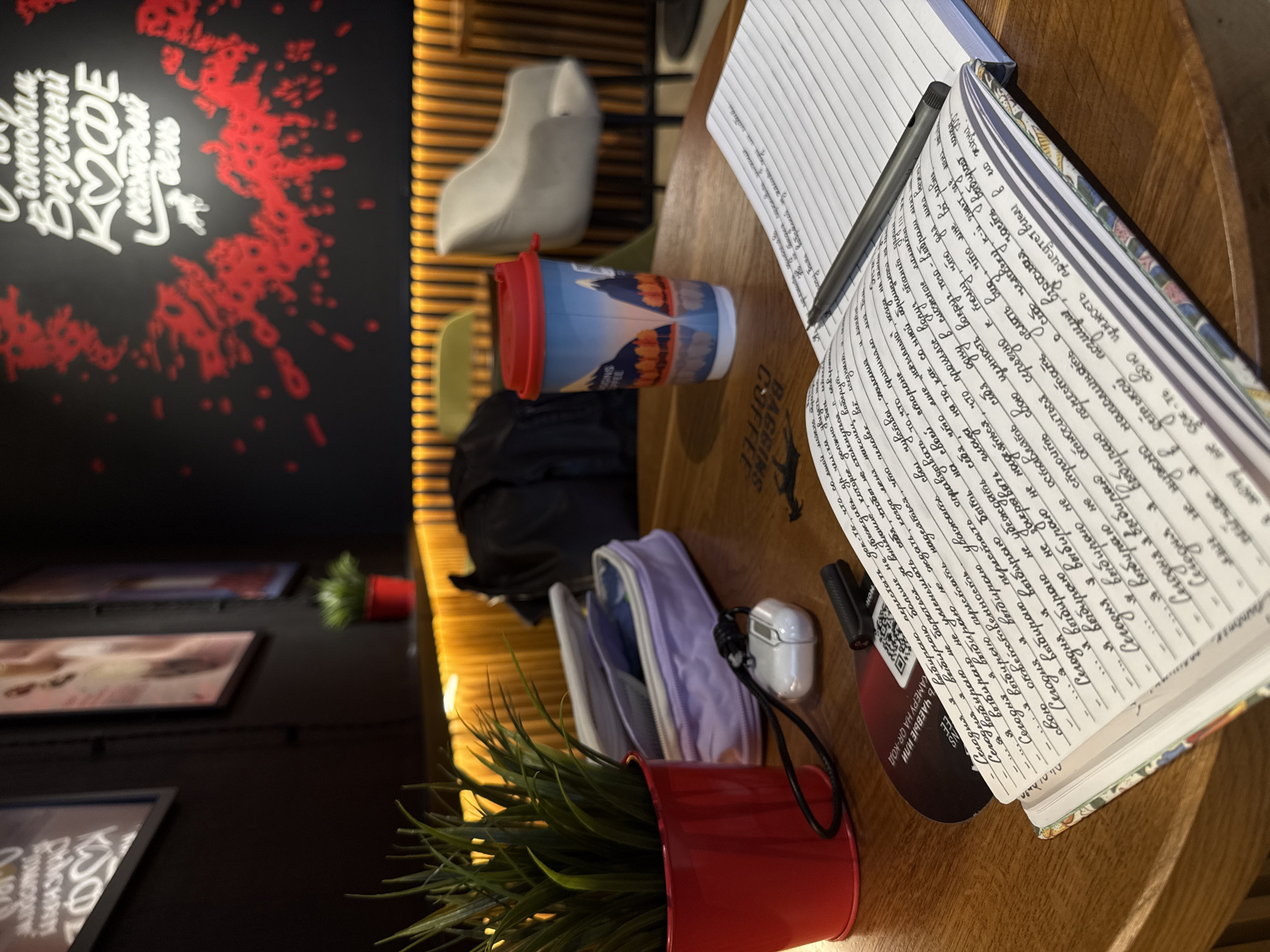 Here's a detailed description of the image:
**General Overview:** The image depicts a table in what appears to be a coffee shop or cafe. There are several items on the table, including notebooks filled with writing, a coffee cup, a pen, a small pot with artificial plants, a pencil case, and earphones. The overall lighting suggests it's daytime.
**Setting:** The presence of a coffee cup and the type of items on the table imply that this is likely a cafe in Saint Petersburg, Russia. In the background, a black wall has a bright, stylized text advertisement written in Russian, which hints at the cafe's name and special offers. The overall style suggests a modern cafe ambiance.
**Objects:**
* **Notebooks:** Two notebooks are visible. One is filled with handwritten text in Russian. The other appears to have blank lined pages.
* **Coffee Cup:** A reusable coffee cup with a red lid.
* **Writing Tools:** A pen rests on one of the notebooks.
* **Plant Pot:** A small red pot contains artificial plants.
* **Pencil Case:** A light purple pencil case.
* **Earphones:** A pair of white wireless earphones.
* **Other:** Other objects, such as a black bag, are partially visible.
**Actions/Interactions:** The presence of notebooks and writing implements suggests someone was or is engaged in writing or studying. The coffee cup indicates someone was drinking a beverage.
**Environment:** The background reveals a contemporary interior design, with dark walls, wooden vertical slat partitions and framed art. There are cafe chairs visible behind the items on the table.
**Overall Impression:** The scene suggests a student or writer spending time in a cafe, possibly studying, journaling, or working on a project. It could also be a casual meeting place.