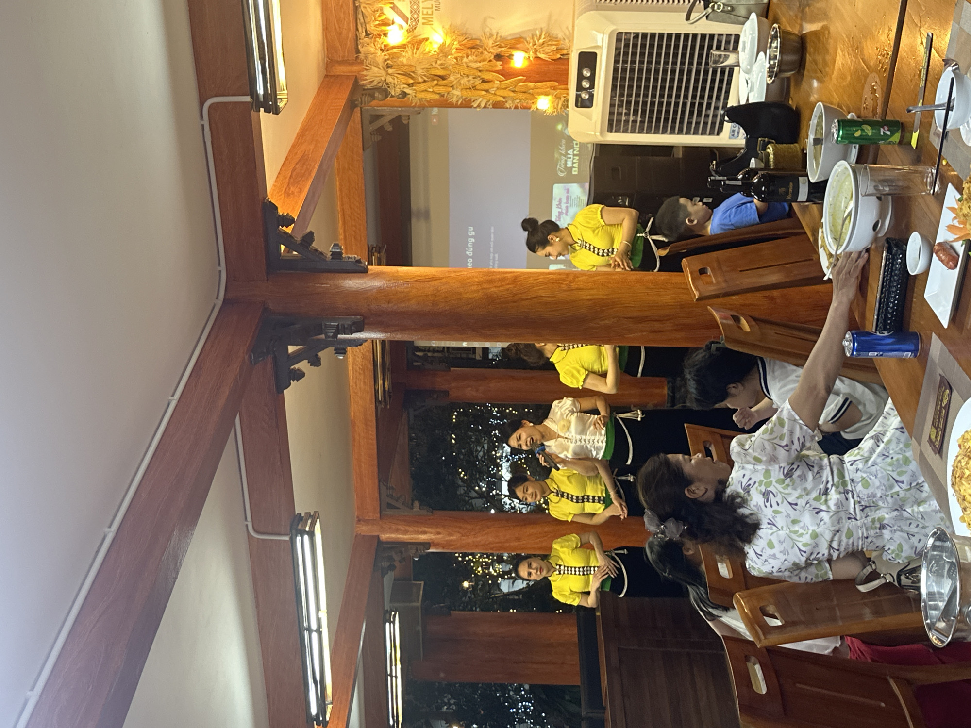 The image depicts an indoor restaurant interior in Nghia Lo Ward, Vietnam, featuring a performance area and a dining table. The ceiling is white with exposed white electrical conduit running along light reddish-brown wooden beams and pillars that structure the space. Rectangular fluorescent light fixtures with dark wooden frames are mounted on the ceiling.
In the background, five women, uniformly dressed in yellow short-sleeved tops with black and white patterned trim, black skirts, and green waist sashes with white tassels, are standing in a line. The woman positioned second from the right holds a microphone and appears to be speaking or singing. Behind them, through large openings, a dark background with numerous small, white decorative lights is visible, suggesting an outdoor or illuminated internal garden area. A white projection screen on the far right wall displays Vietnamese text, including "theo đúng gu" and "trong khuôn MÙA BẢN NỖ." To the right of the screen, a wall is decorated with numerous dried corn cobs intertwined with warm-toned string lights. A white oscillating air cooler stands on the floor in the foreground-right.
In the foreground, a large, dark wooden dining table is occupied by diners. A woman with dark hair, wearing a white dress with a green and purple floral pattern, is seated with her back partially to the camera, looking towards a young boy seated to her right, who is wearing a white shirt with dark stripes. Another young boy in a blue t-shirt is visible further to the right at the table. The table is set with various white bowls and plates containing food, including what appears to be a noodle or rice dish and a sausage. Beverages present include a blue soda can, a green 7UP can, and a dark glass bottle labeled "Verano" (wine). A silver ice bucket, clear drinking glasses, and a black rectangular object (possibly a remote control) are also on the table. A person's hand is visible reaching towards a dish on the table.