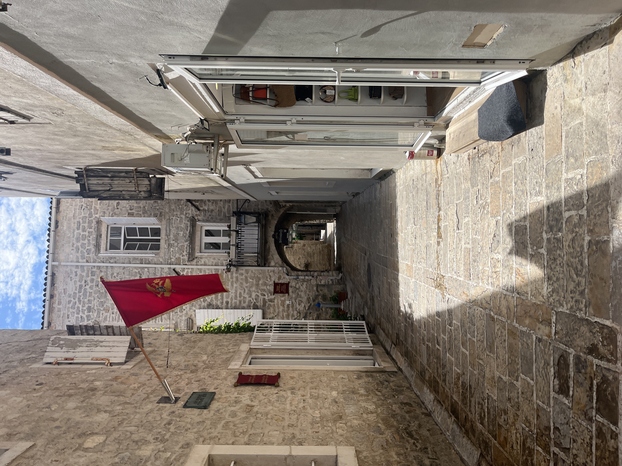 The image depicts a narrow, cobblestone alleyway in the Old Town of Budva, Montenegro. The alley is lined with stone buildings, some with wooden shutters and balconies. A red flag with a golden eagle emblem, likely the flag of Montenegro, hangs from a pole attached to one of the buildings on the left. On the right side of the alley, a shop with large glass doors displays handbags on shelves. The ground is paved with irregular stone blocks, and sunlight casts long shadows across the alley. The sky above is partly cloudy. The overall scene conveys a sense of historic European architecture and a bright, sunny day.