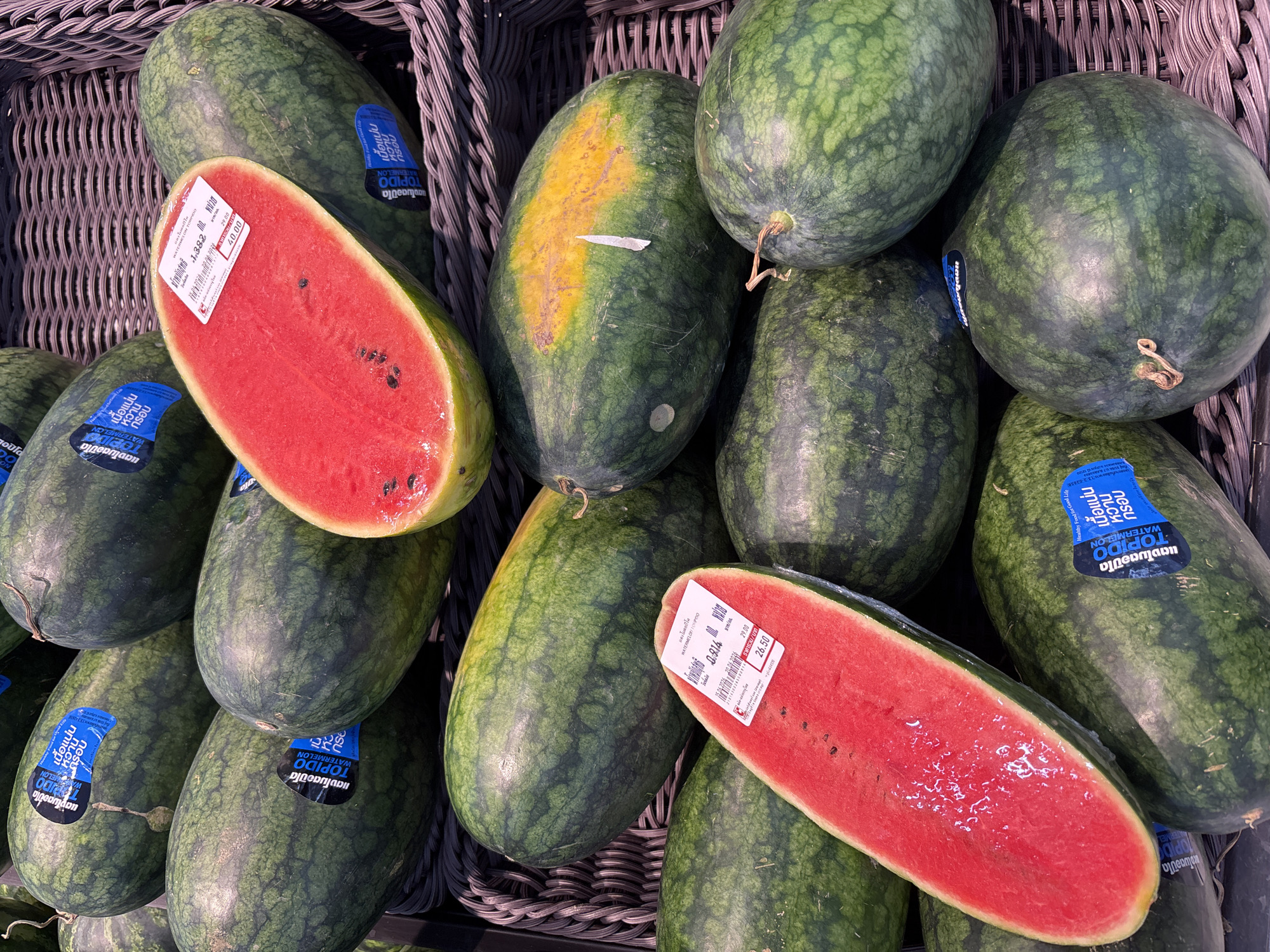 The image shows a display of watermelons at a market in Ban Bang Rak, Thailand. Several whole watermelons are visible, some with blue stickers indicating the "TOPIDO WATERMELON" brand. One watermelon is cut in half, revealing its red flesh and seeds. Another half-watermelon is displayed separately, also showing its vibrant red interior. Price tags are attached to some of the watermelons. The watermelons are arranged in woven baskets. The environment appears to be an indoor market or grocery store setting.