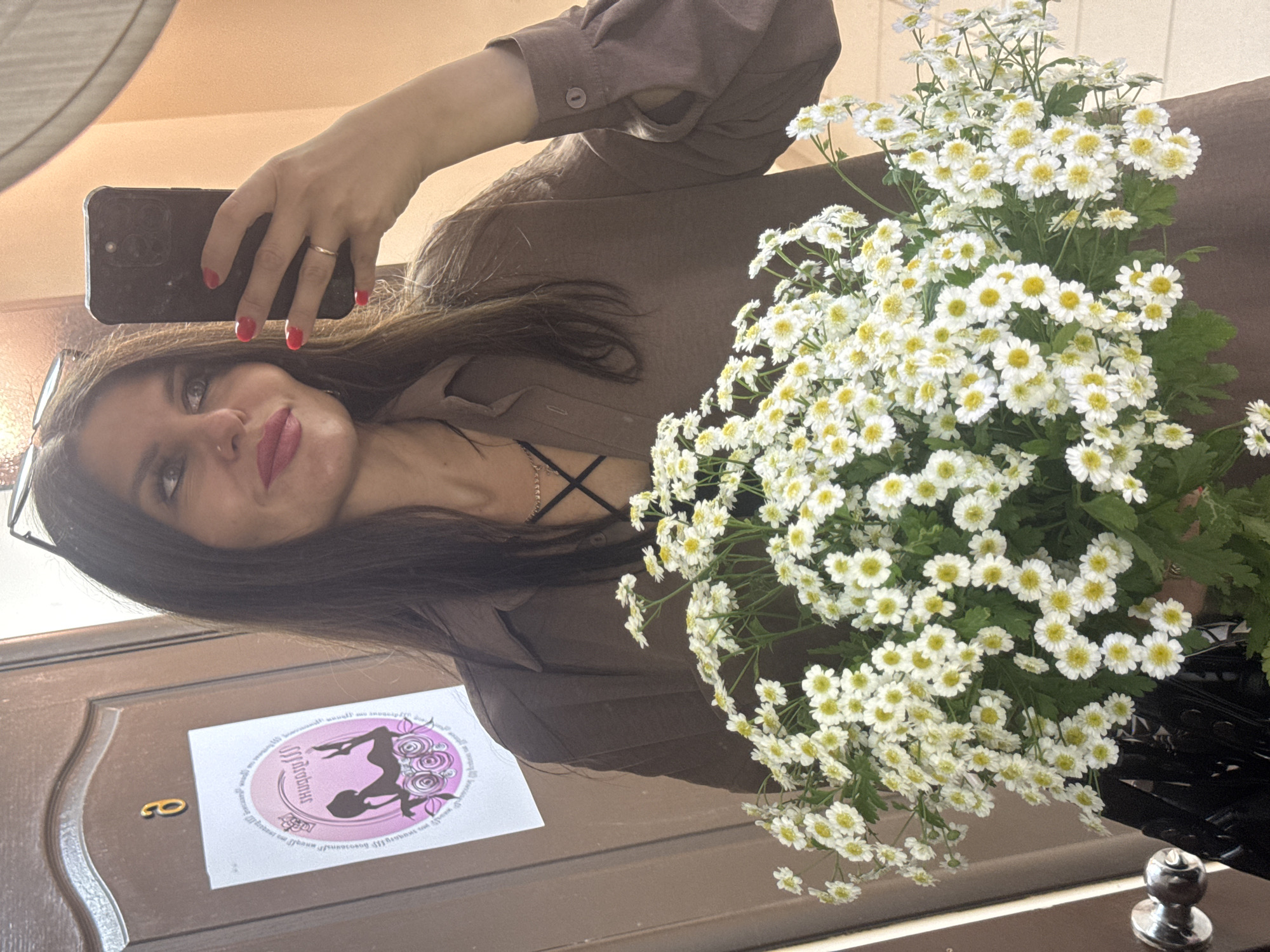 A woman is taking a mirror selfie in what appears to be an apartment or hallway. She is holding a smartphone in her left hand, with her nails painted red. She is wearing a brown dress. A large bouquet of white flowers with yellow centers is held in front of her, obscuring much of her body. The flowers are brightly lit, suggesting good lighting conditions, possibly daytime.
In the mirror's reflection, a sticker is visible on a door. The sticker is oval-shaped and pink, featuring a silhouette of a woman with flowers. Some text is present around the edges of the sticker, but it is too blurry to read. A golden numeral "8" is also visible on the door, next to the sticker. The woman's face is visible in the reflection, and she is smiling slightly. She has dark hair, which falls over her shoulders. She is wearing glasses.
The overall impression is a casual self-portrait, possibly taken to capture a moment or to share an outfit or the flowers. The reflection in the mirror reveals a wooden door and a light-colored wall in the background.