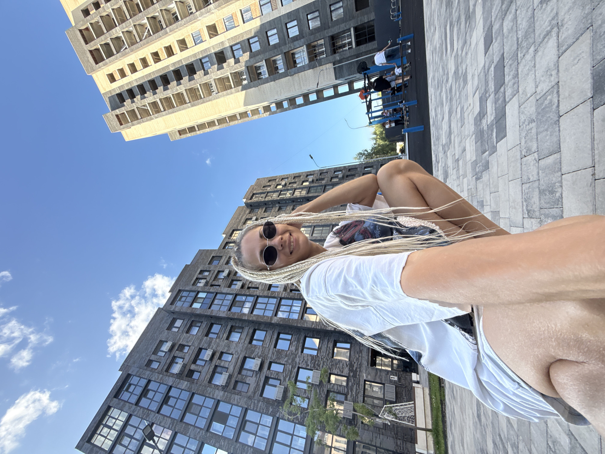 The image depicts a woman with blonde braided hair, wearing sunglasses and a white t-shirt, crouching on a paved area. She is positioned in the foreground, looking towards the camera. Behind her, two multi-story residential buildings are visible, one with a dark brick facade and the other with a lighter, possibly unfinished, exterior. A children's playground with several individuals, including adults and children, is situated between the buildings. The scene appears to be set in an urban environment, possibly a residential complex in Rusanovo, Russia, under a clear blue sky with some scattered clouds. The paving stones are a mix of grey and darker shades, forming a patterned surface.