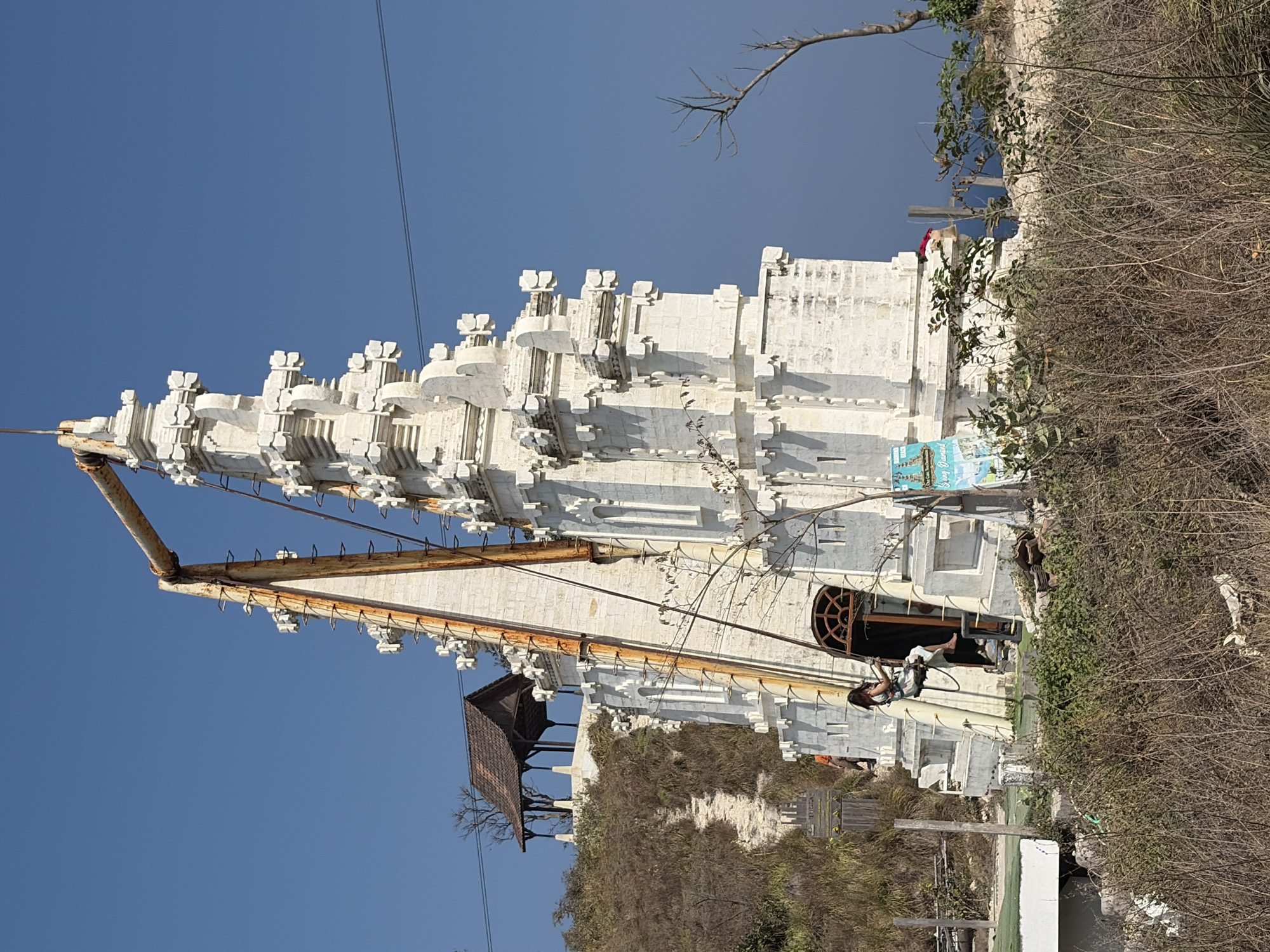 A white, intricately carved tower stands prominently in the foreground, resembling a temple or monument. A large, rusty metal structure arches over the top of the tower, with a thin rope extending down its side, to which a woman is attached, appearing to be on a swing or zipline. The woman is wearing a light-colored outfit and has her legs extended. The tower is surrounded by dry, yellowish grass and some green bushes. To the left of the tower, a path winds up a grassy hill towards a small, dark-roofed pavilion. In the background, the bright blue sky meets the darker blue ocean. A few trees and power lines are visible. The scene is set in Nusa Penida, Indonesia, near a cliff overlooking the sea.