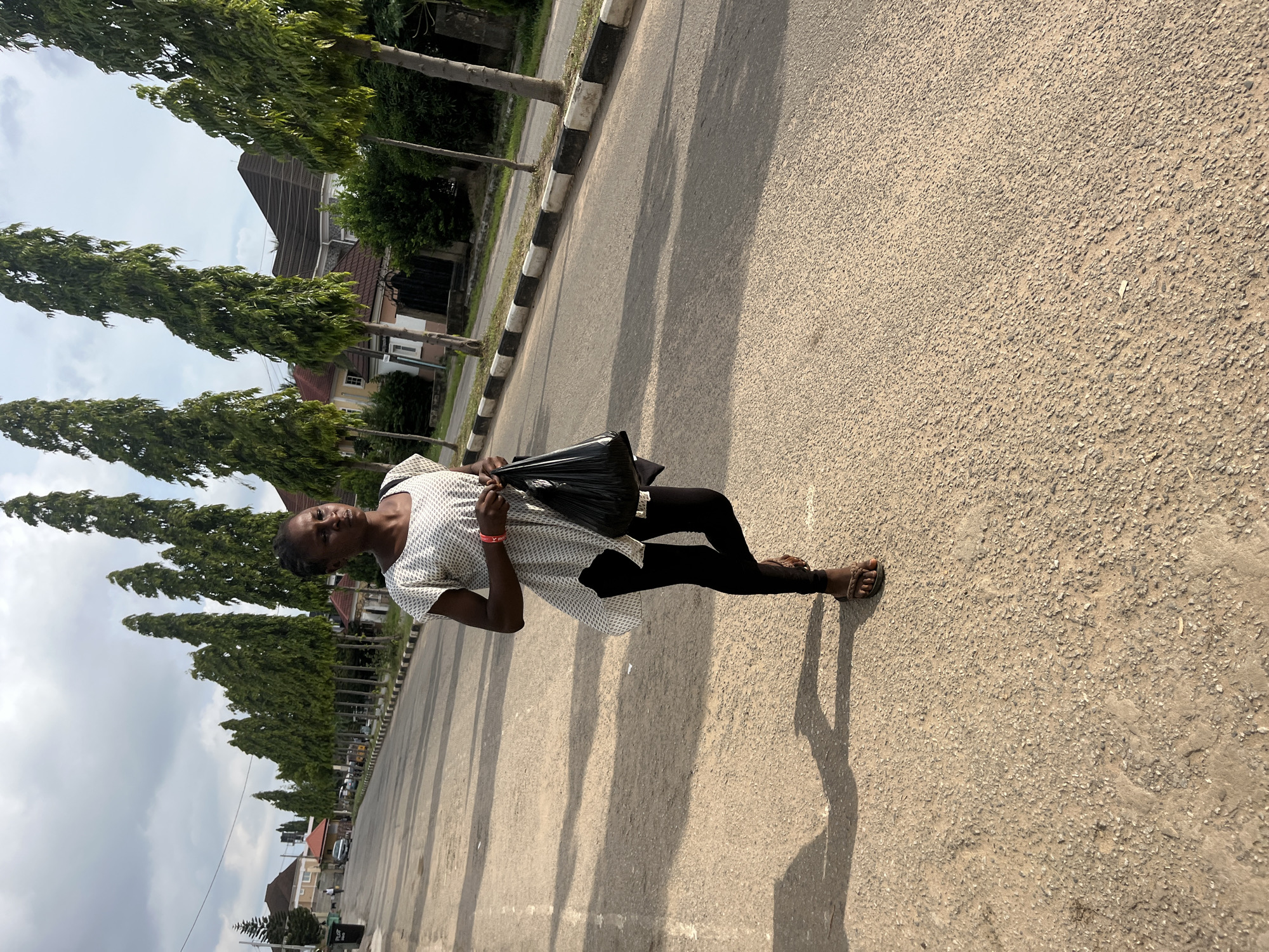 An adult female is depicted walking on a paved street in Abuja, Nigeria. She is wearing a white and patterned tunic over black leggings and brown sandals. She carries a black plastic bag in her right hand. The street is lined with tall, thin trees. Houses with pitched roofs are visible in the background, along with a cloudy sky. The lighting suggests it is daytime.
