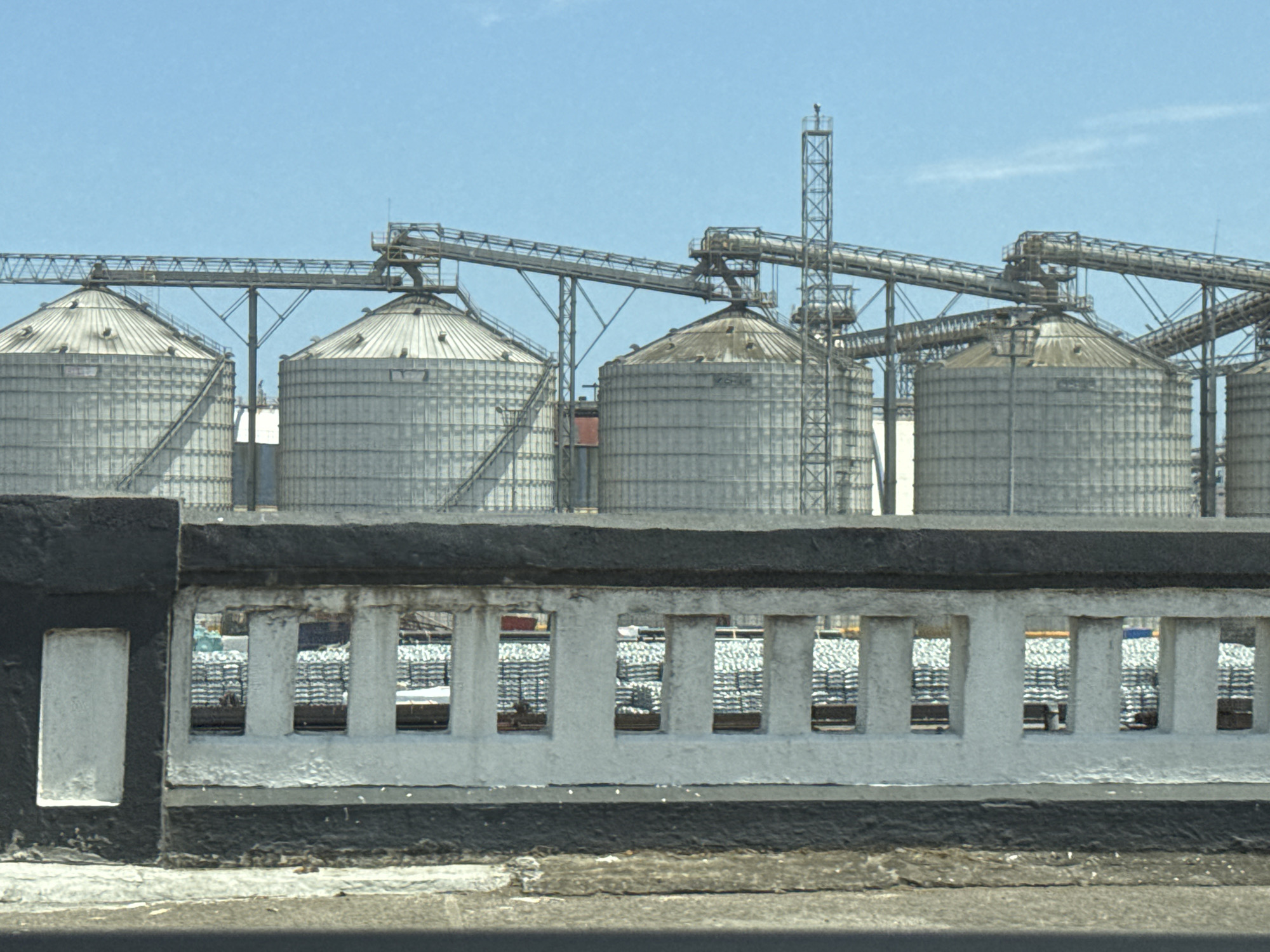 The image shows a row of five large, cylindrical grain silos behind a low, weathered concrete wall with a decorative, repeating rectangular pattern. The silos are made of corrugated metal and have conical roofs. Overhead, a series of metal conveyor belts and support structures connect the silos, extending towards a taller metal tower structure in the background. Beyond the wall, stacked bags, possibly containing grain, are visible. The sky is clear and blue. The location is likely Veracruz City, Mexico, given the industrial infrastructure and architectural style.