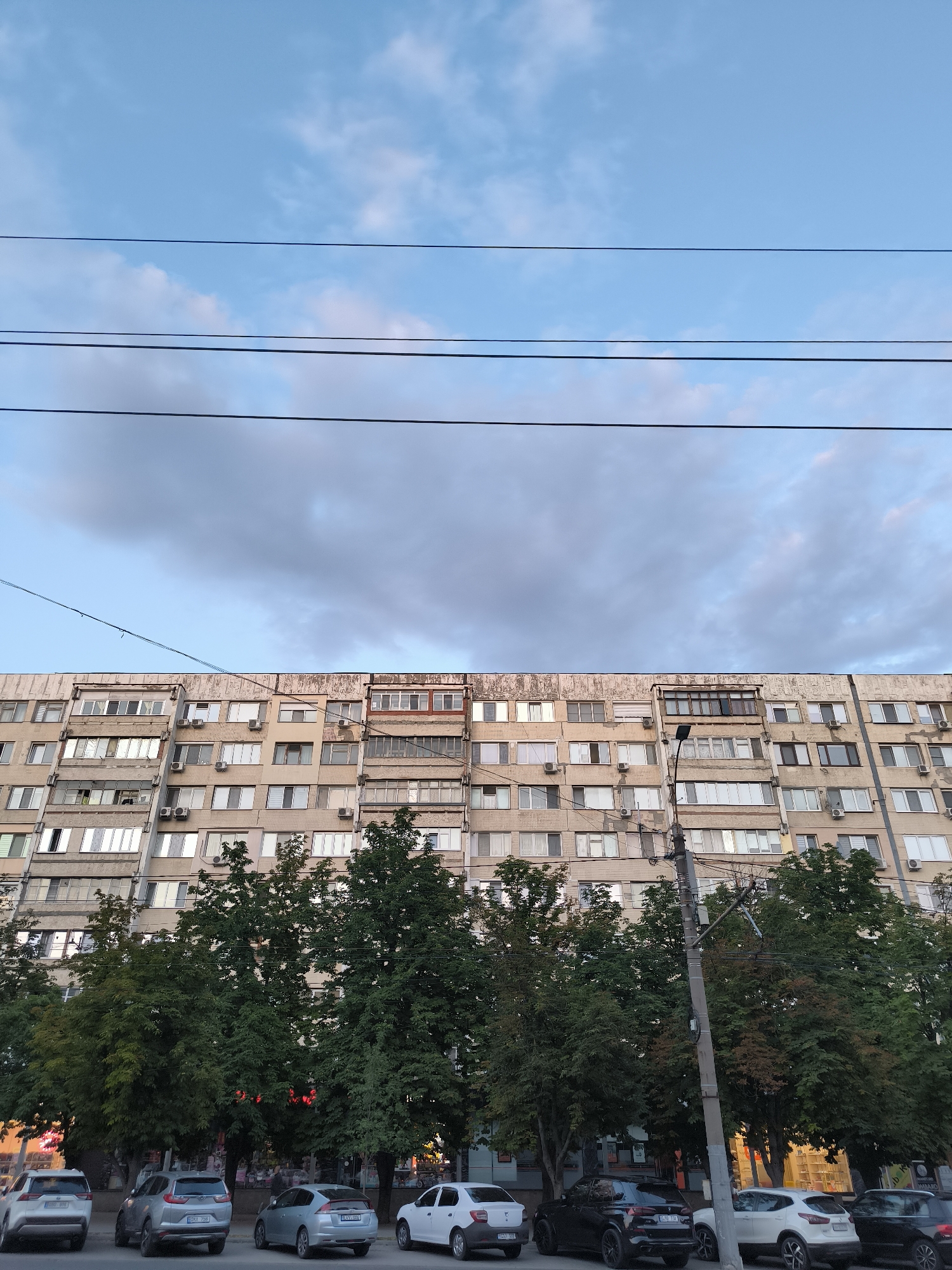 The image shows a large, multi-story apartment building in Chișinău, Moldova. The building is made of concrete and has many windows and balconies. There are trees in front of the building and several cars parked on the street. Overhead, there are power lines against a sky with clouds. The overall scene depicts a typical urban environment in Chișinău.