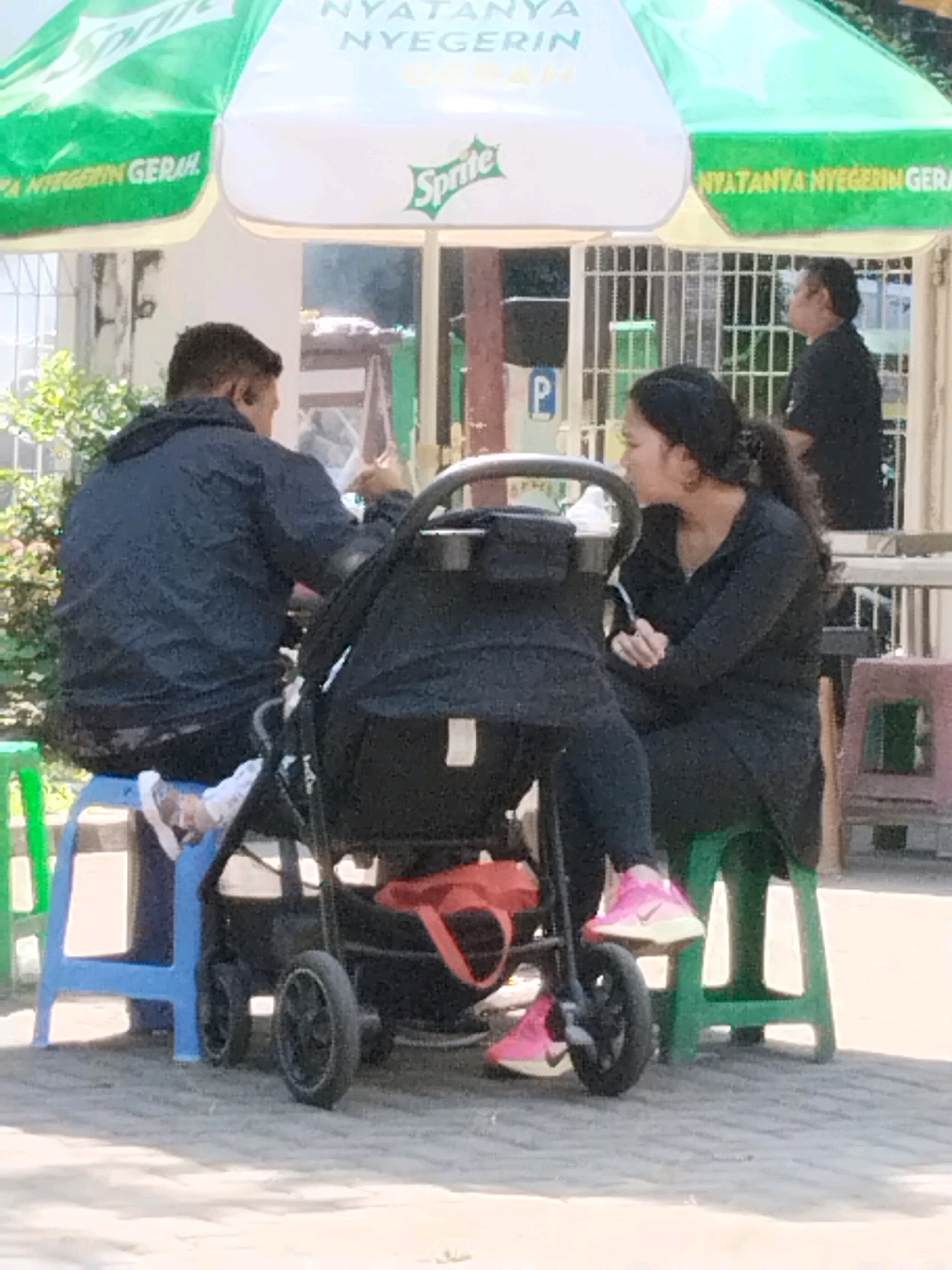 The image depicts a street scene in Surabaya, Indonesia, likely near a food stall or outdoor seating area. Two adults and a baby in a stroller are visible. The man on the left, wearing a dark blue hooded jacket and black pants, is seated on a blue plastic stool, facing away from the camera. He appears to be smoking. A woman on the right, dressed in a black outfit, is seated on a green plastic stool, facing the man and the stroller. She is looking towards the man and has her hands clasped. Between them is a black baby stroller. In the background, a Sprite umbrella provides shade. Another person in a black shirt is visible in the distance, possibly a vendor or another customer. The ground is paved with light-colored stones. The overall impression is of a casual, everyday scene in a public space.